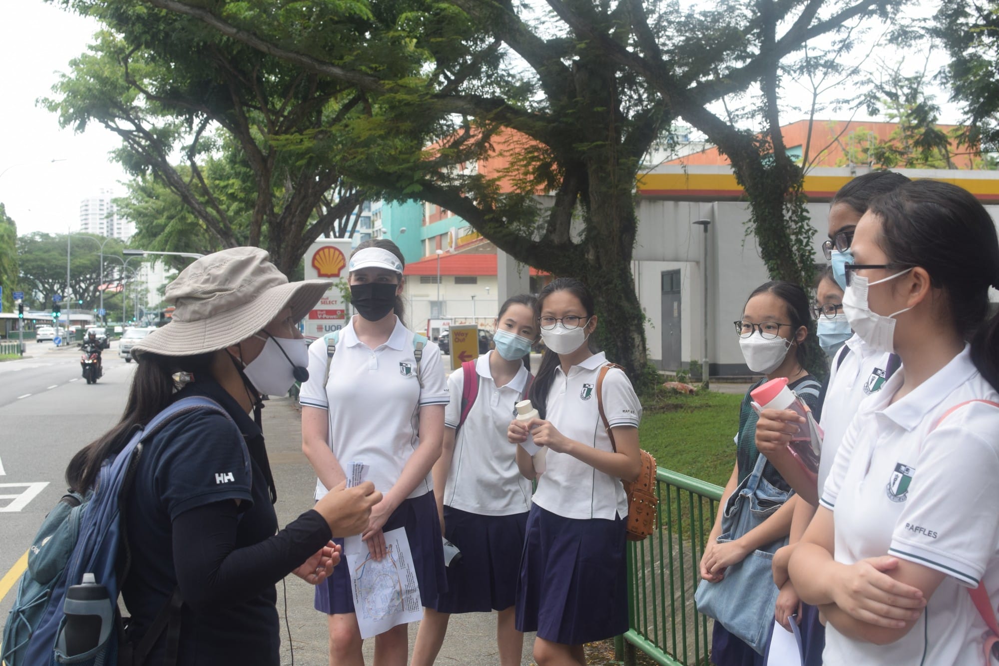 A masked guide wearing a hat speaks to masked students in white uniforms on a city sidewalk near a Shell station.