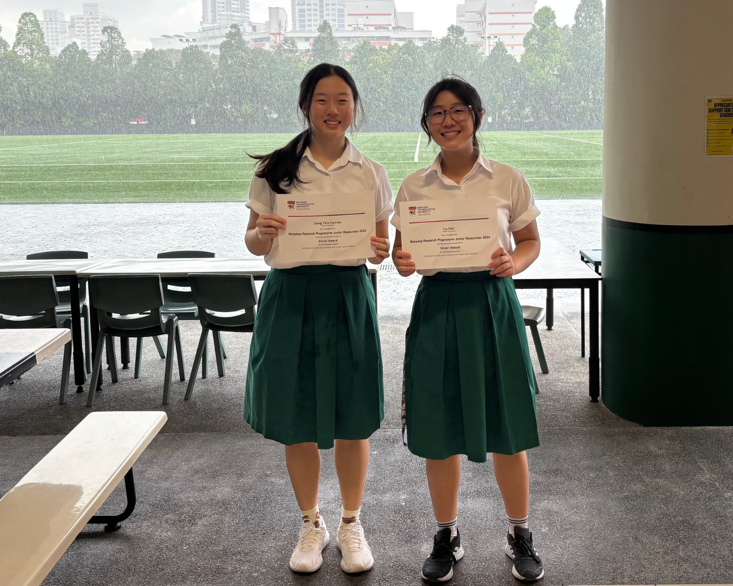 Two smiling schoolgirls in white shirts and green skirts hold Silver Award certificates indoors with a rainy field behind them.