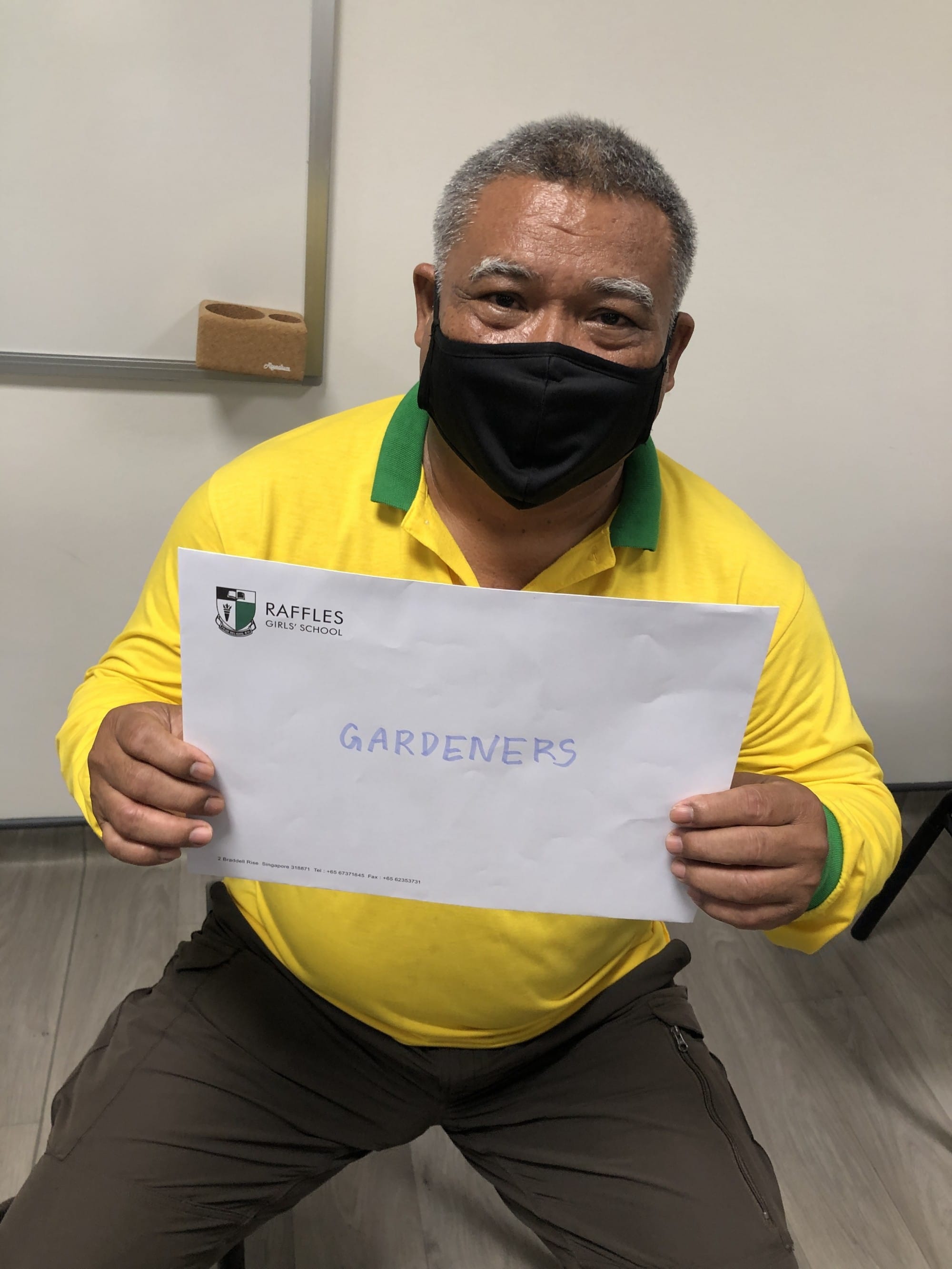 Man in yellow shirt and black mask holds a Raffles Girls' School paper reading "GARDENERS."