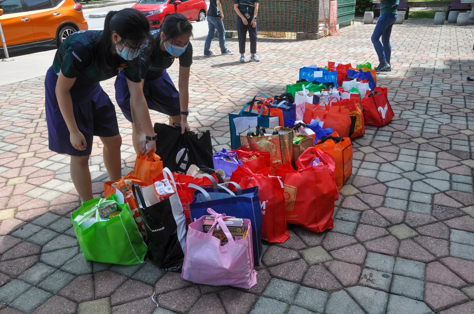 Two masked girls arrange many colorful reusable bags filled with goods on a paved outdoor area.
