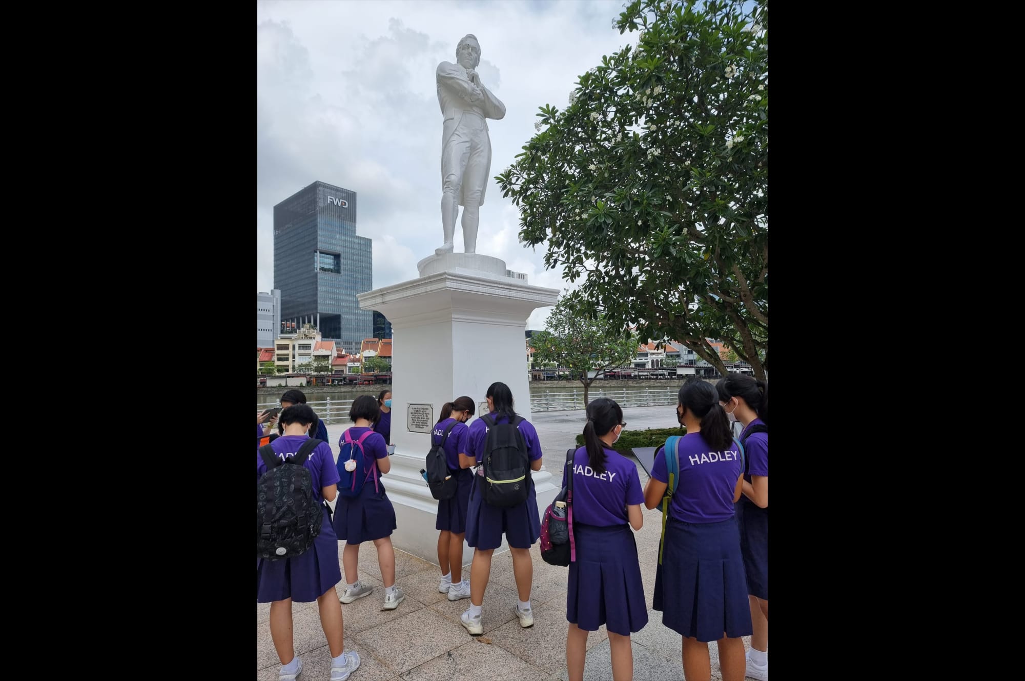 Students in purple Hadley uniforms stand before a tall white statue near a river with city buildings behind.