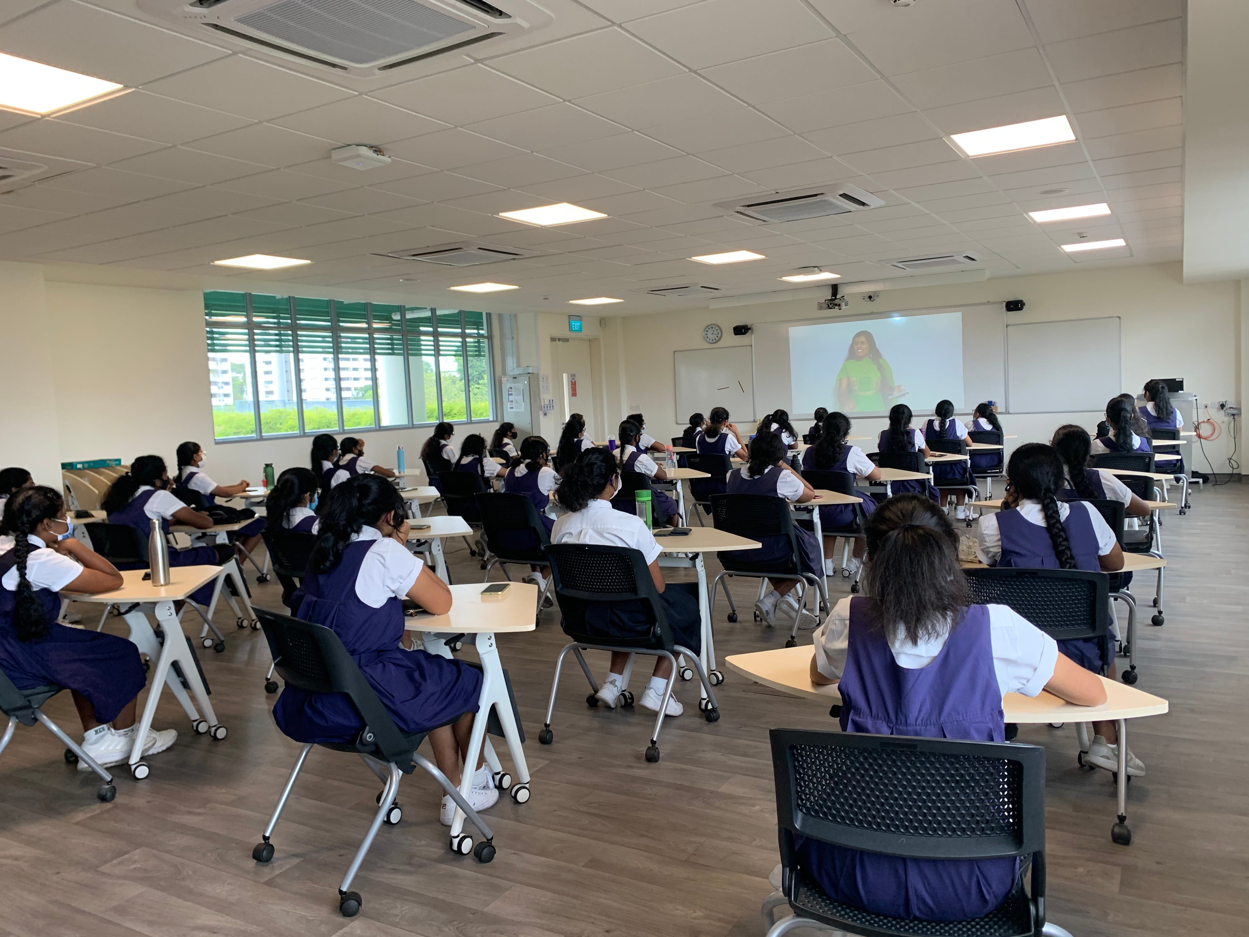 Students in navy and white uniforms sit at desks in a bright classroom watching a projected video on a screen.