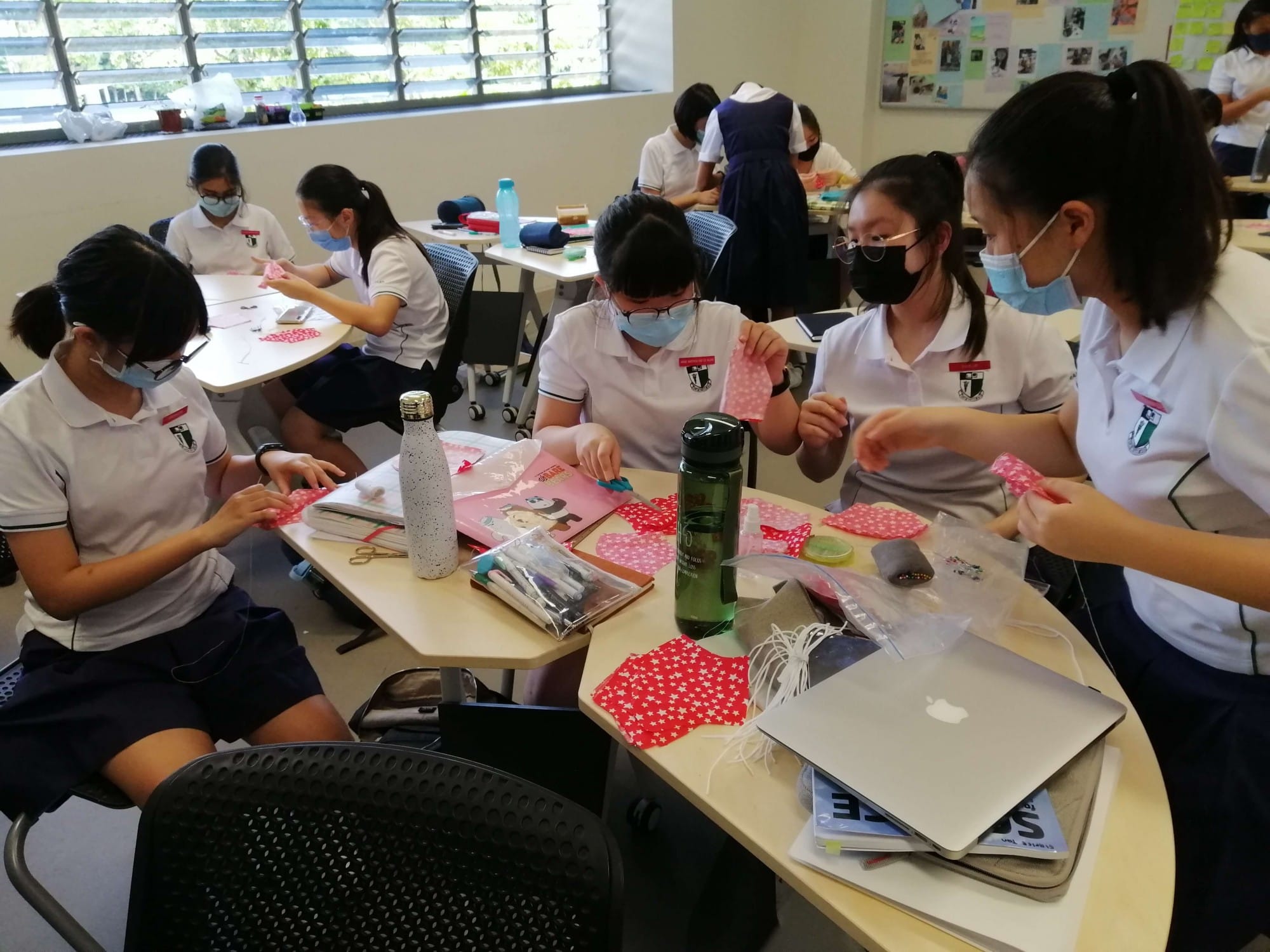 Students in white uniforms and face masks work on a fabric crafting activity at classroom desks.