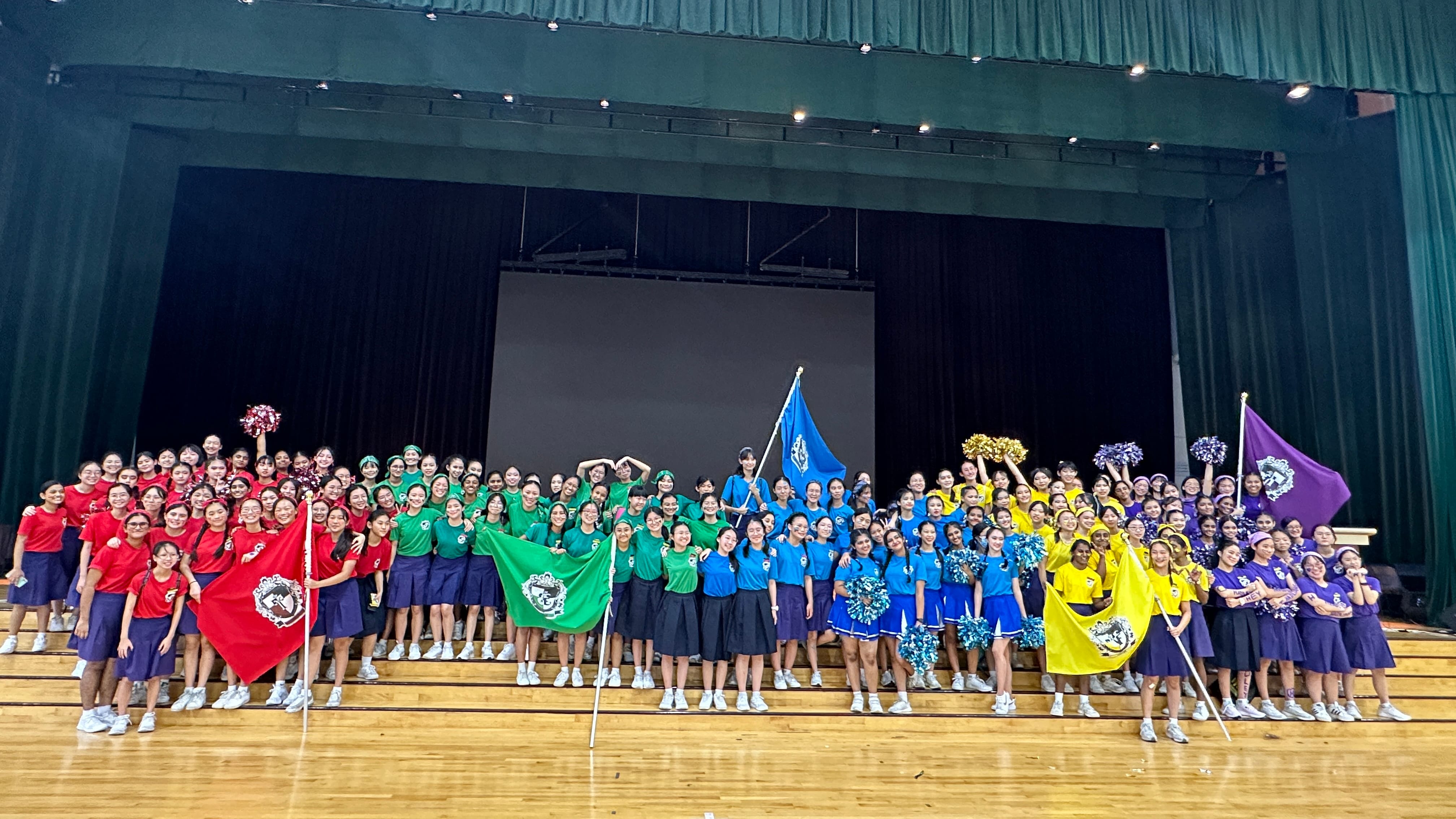 Large group of students in red, green, blue, yellow, and purple shirts holding colored flags in a school hall.