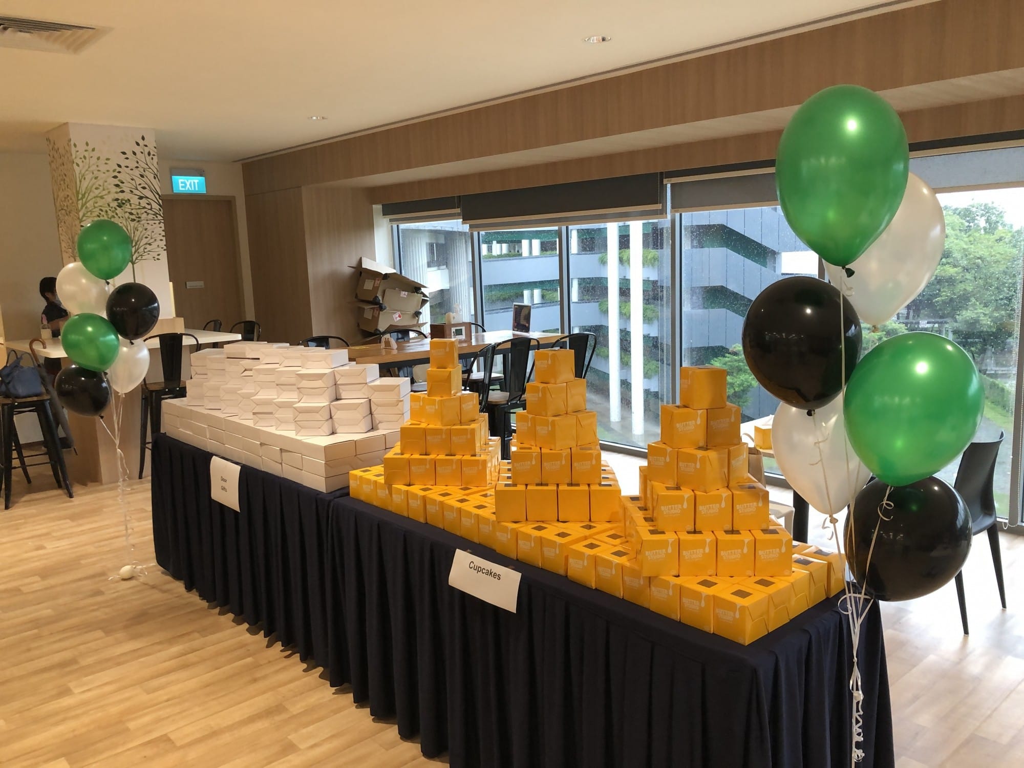 A table stacked with yellow cupcake boxes and white boxes, flanked by green, black, and white balloons in a bright room.