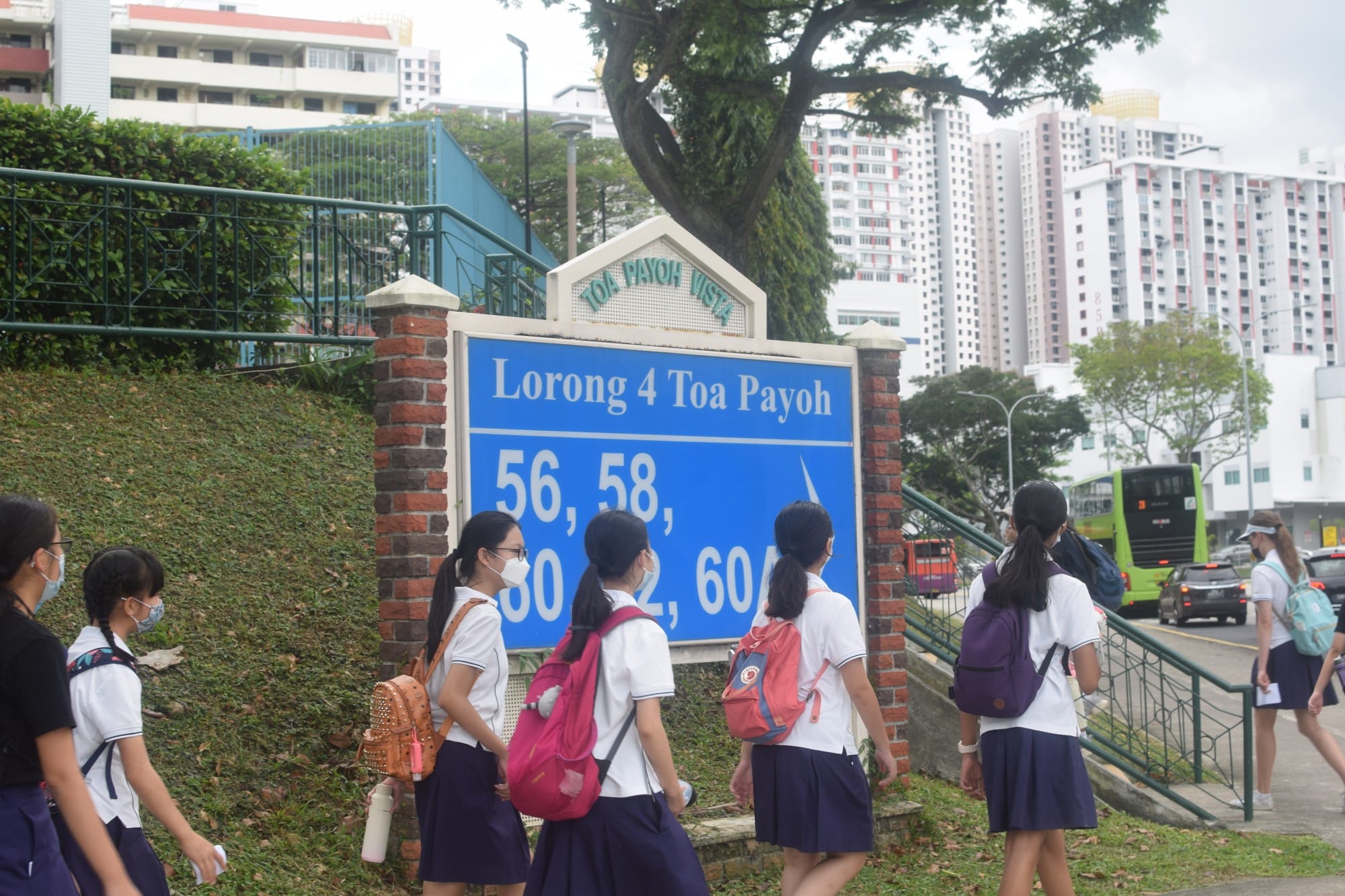 Students in white uniforms and dark skirts walk past a blue street sign reading Lorong 4 Toa Payoh in Singapore.