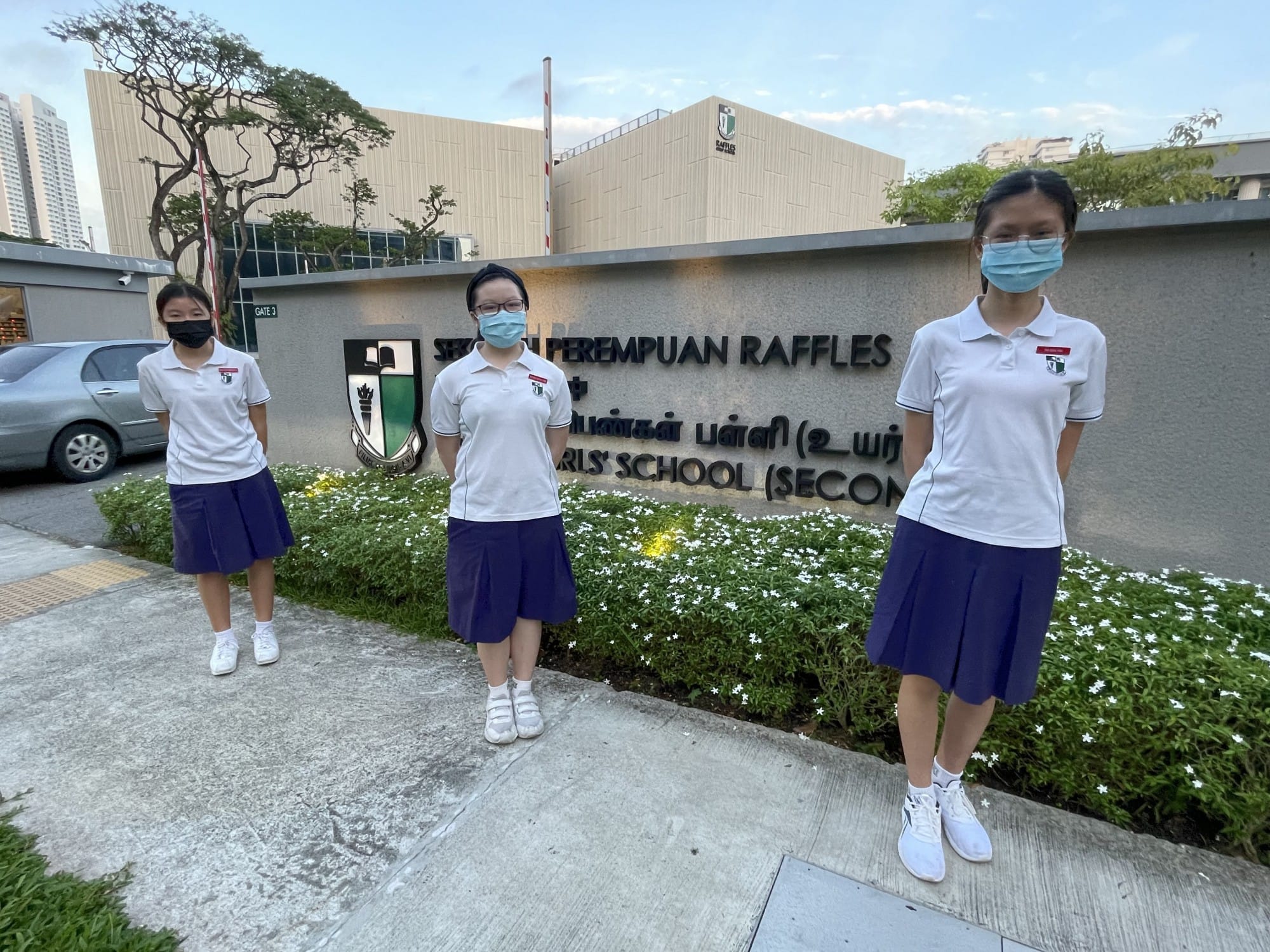 Three masked schoolgirls in white shirts and blue skirts stand before the Raffles Girls School sign.