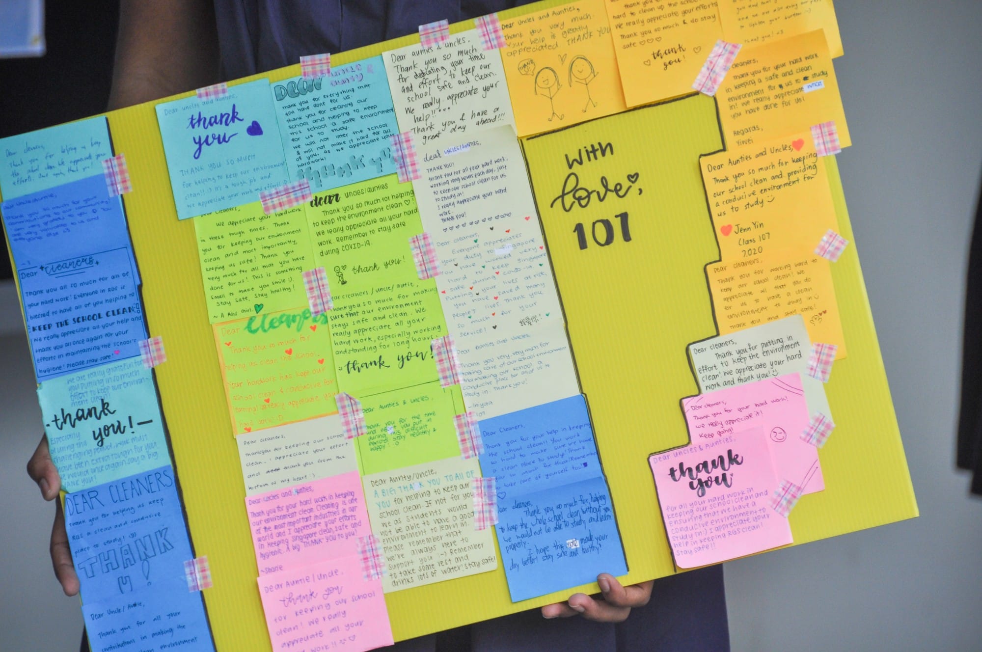 A person holds a yellow board covered with colorful thank-you notes addressed to school cleaners, signed "With love, 101."