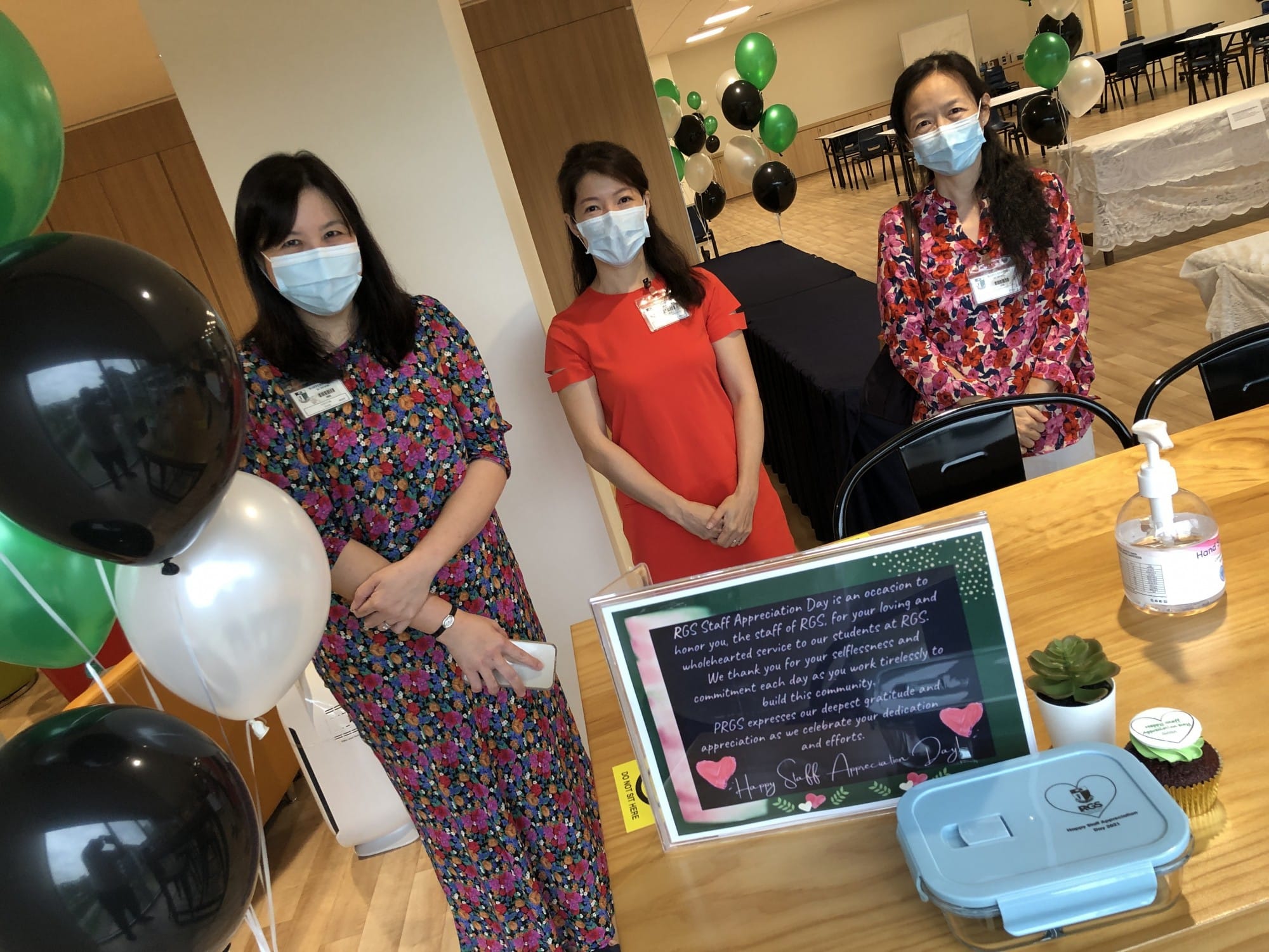 Three masked women stand near a table with a Happy Staff Appreciation Day sign, balloons, and hand sanitizer.