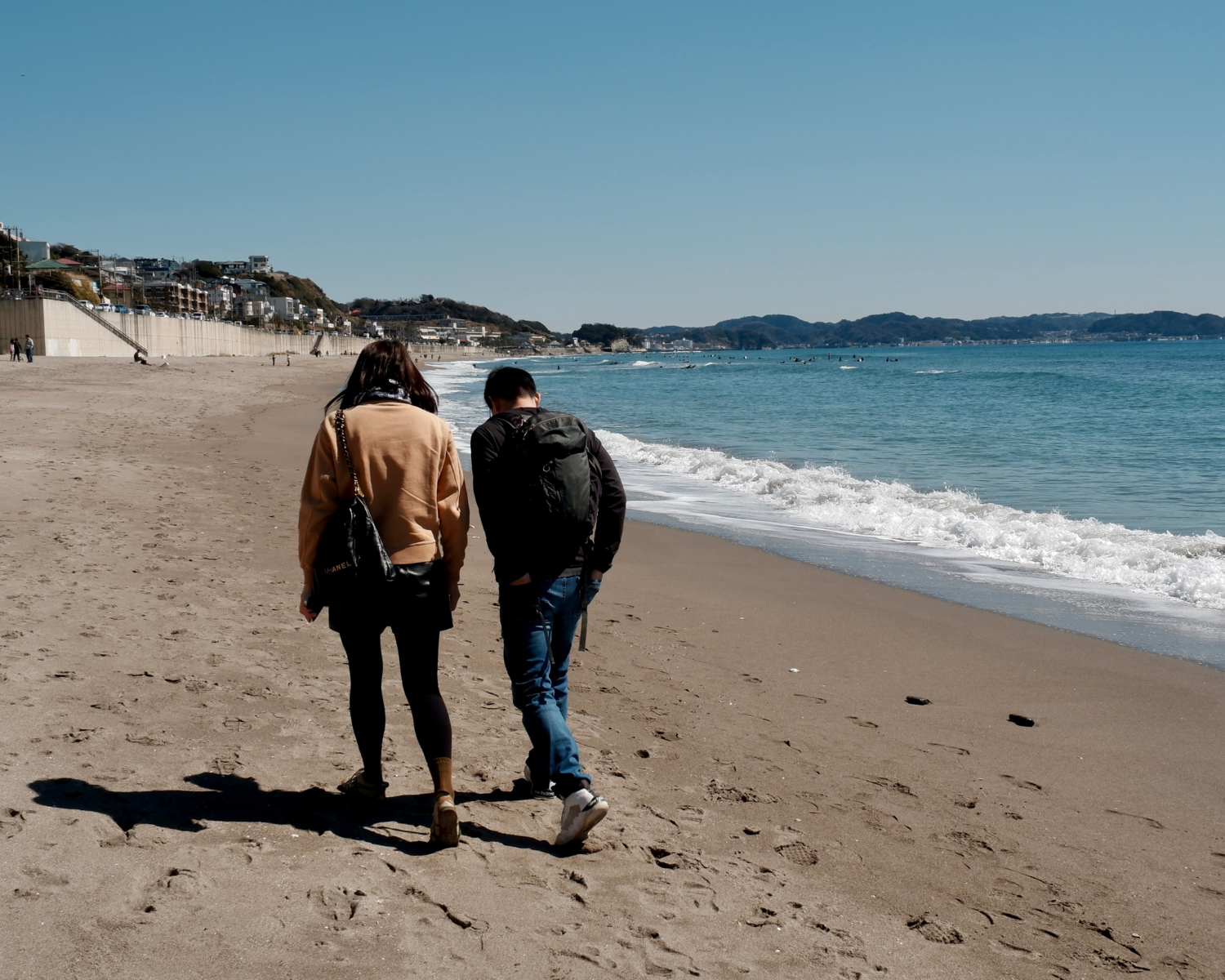 Two people walking along a sandy beach with ocean waves, hills, and buildings visible in the background.