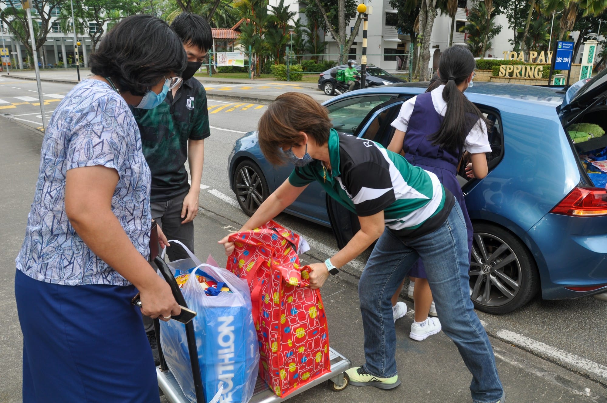 Four masked people unload colorful bags from a blue car onto a trolley on a Singapore street.
