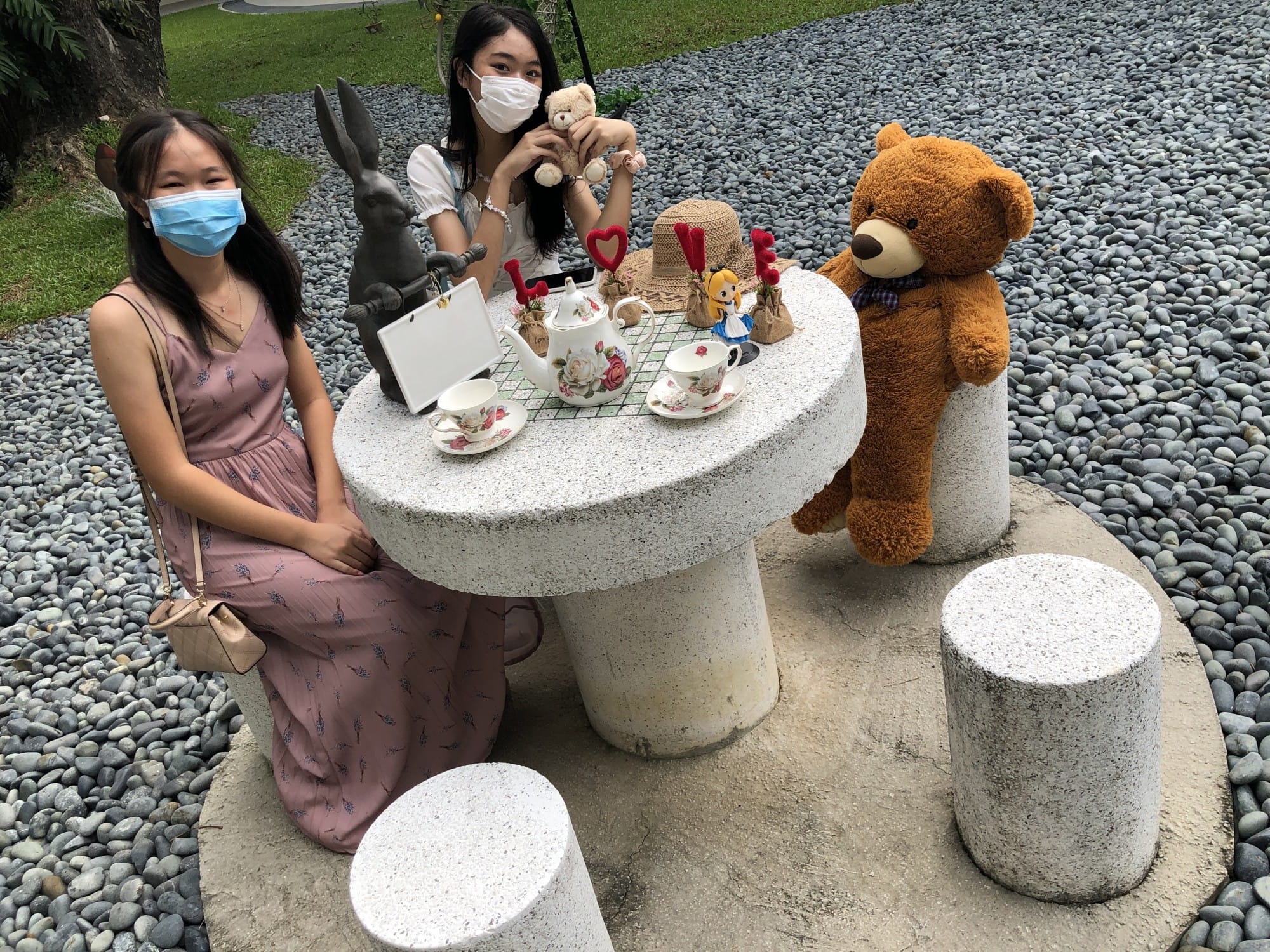 Two masked women sit at a stone outdoor table with a tea set, large teddy bear, and rabbit statue on a pebble ground.