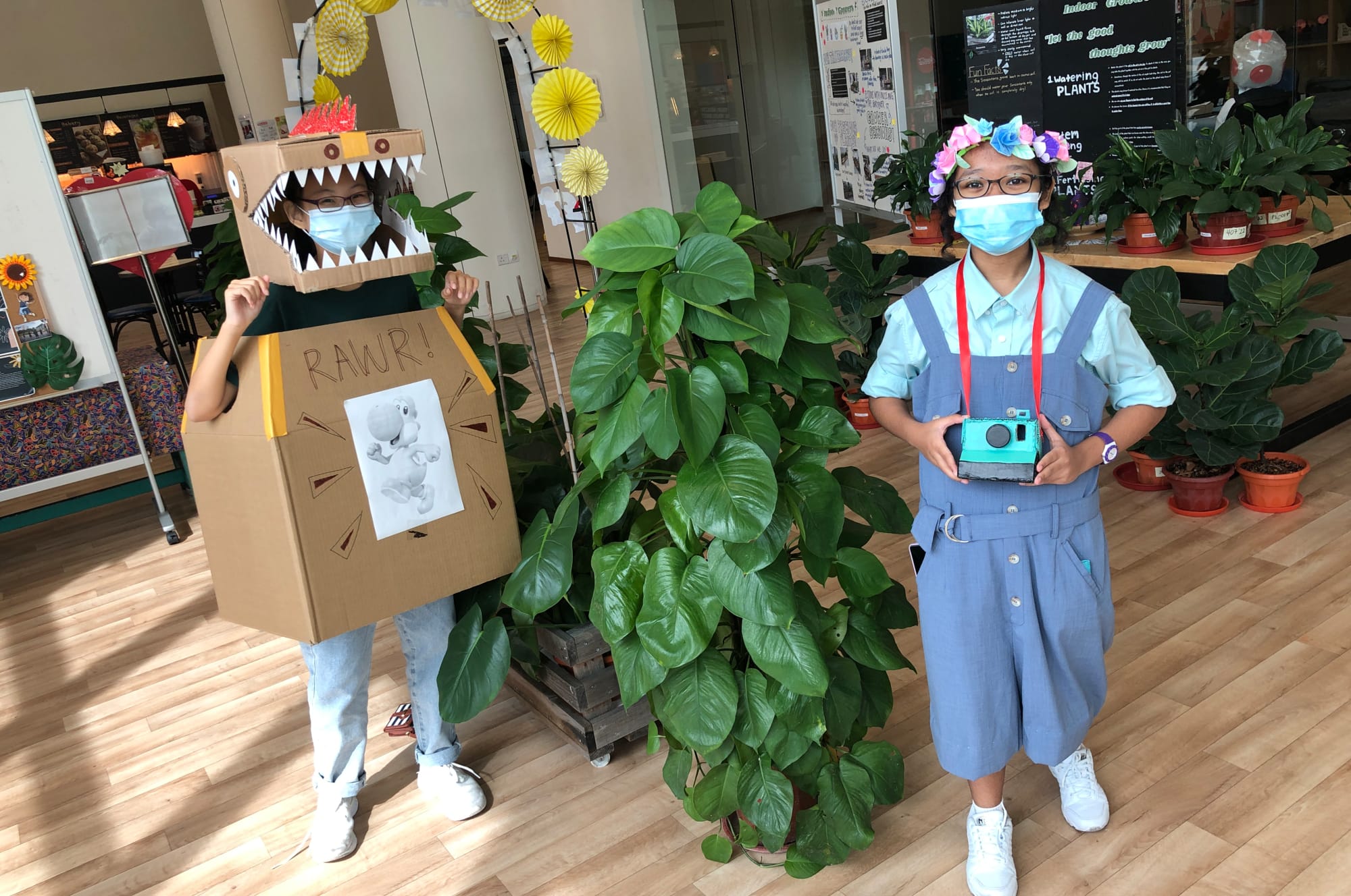 Two masked kids indoors by plants; one in a cardboard dinosaur costume, the other with flower crown holds camera.