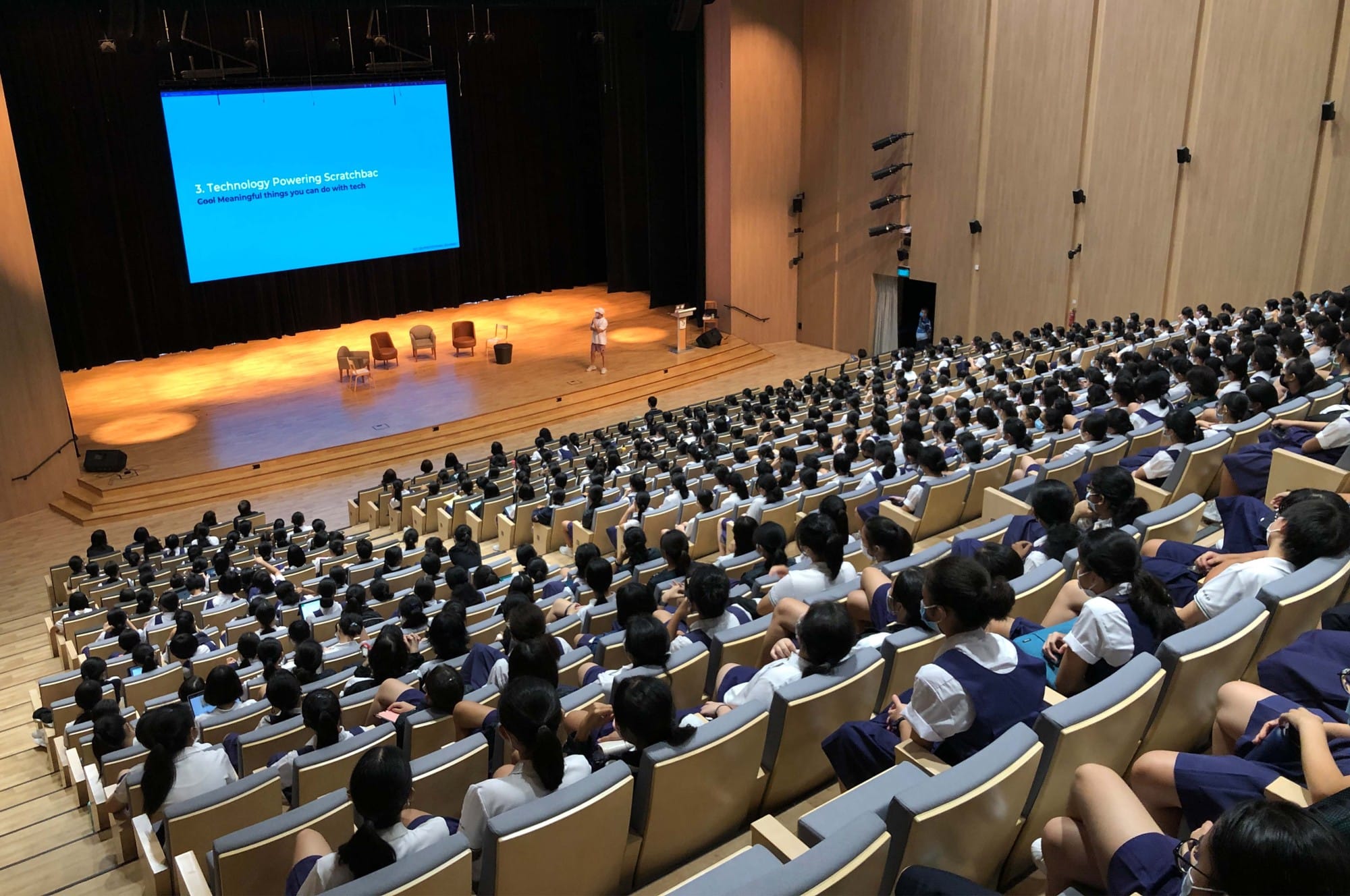 A large auditorium filled with uniformed students watching a speaker on stage with a blue presentation slide.