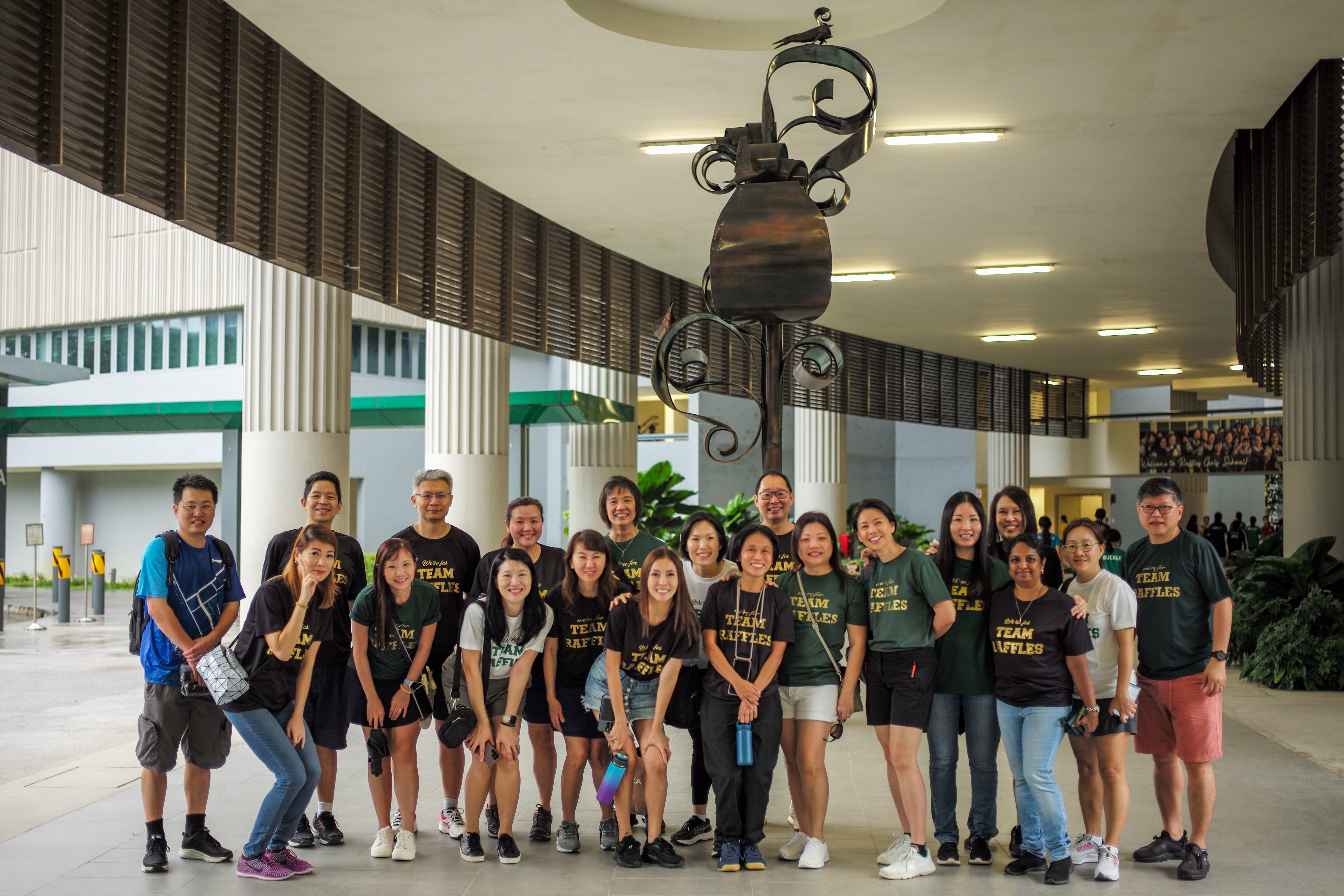 Group of about 20 people in dark green Team Raffles shirts posing under a metal sculpture in a covered corridor.