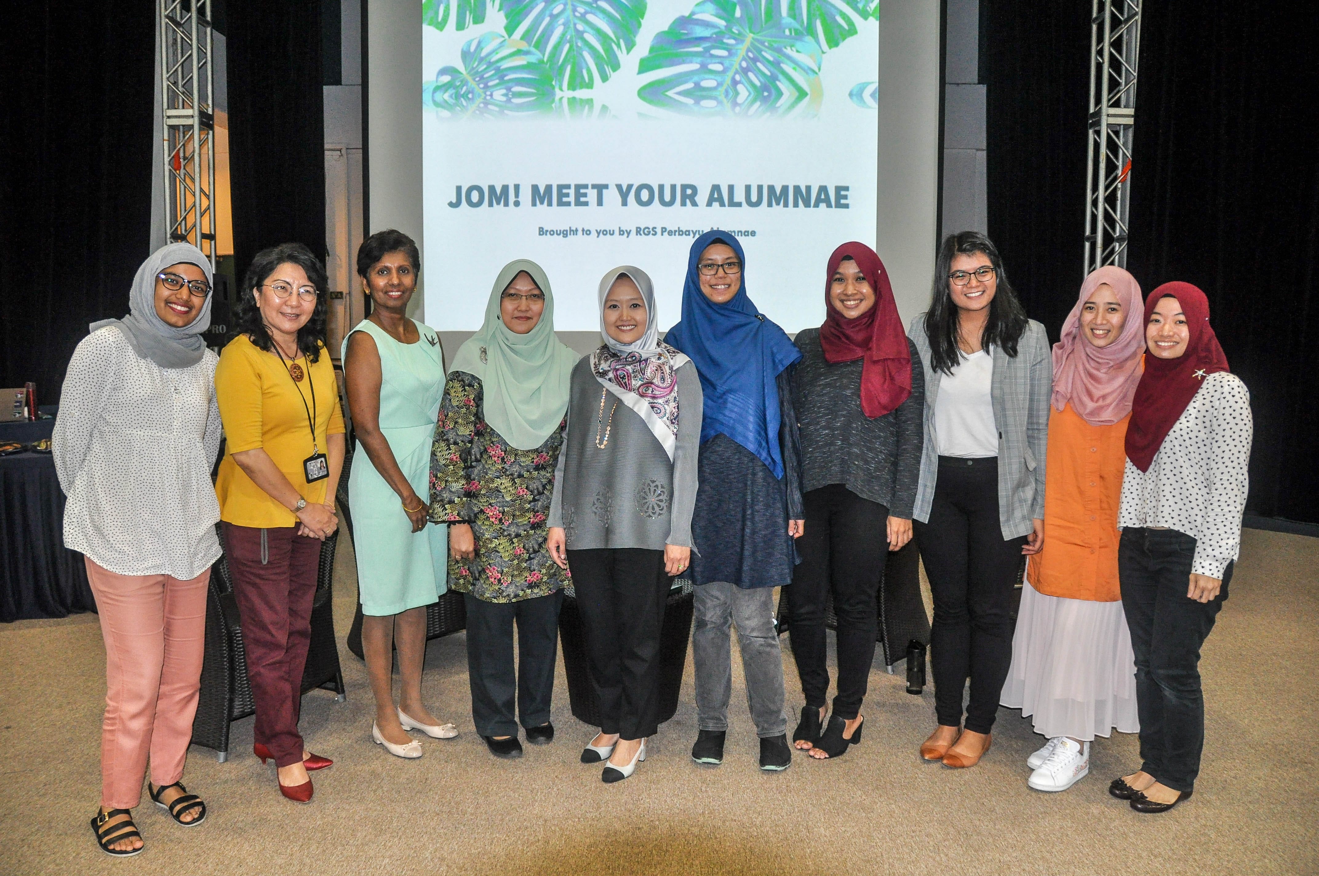 Ten women stand smiling in front of a screen reading "JOM! MEET YOUR ALUMNAE" at an event.