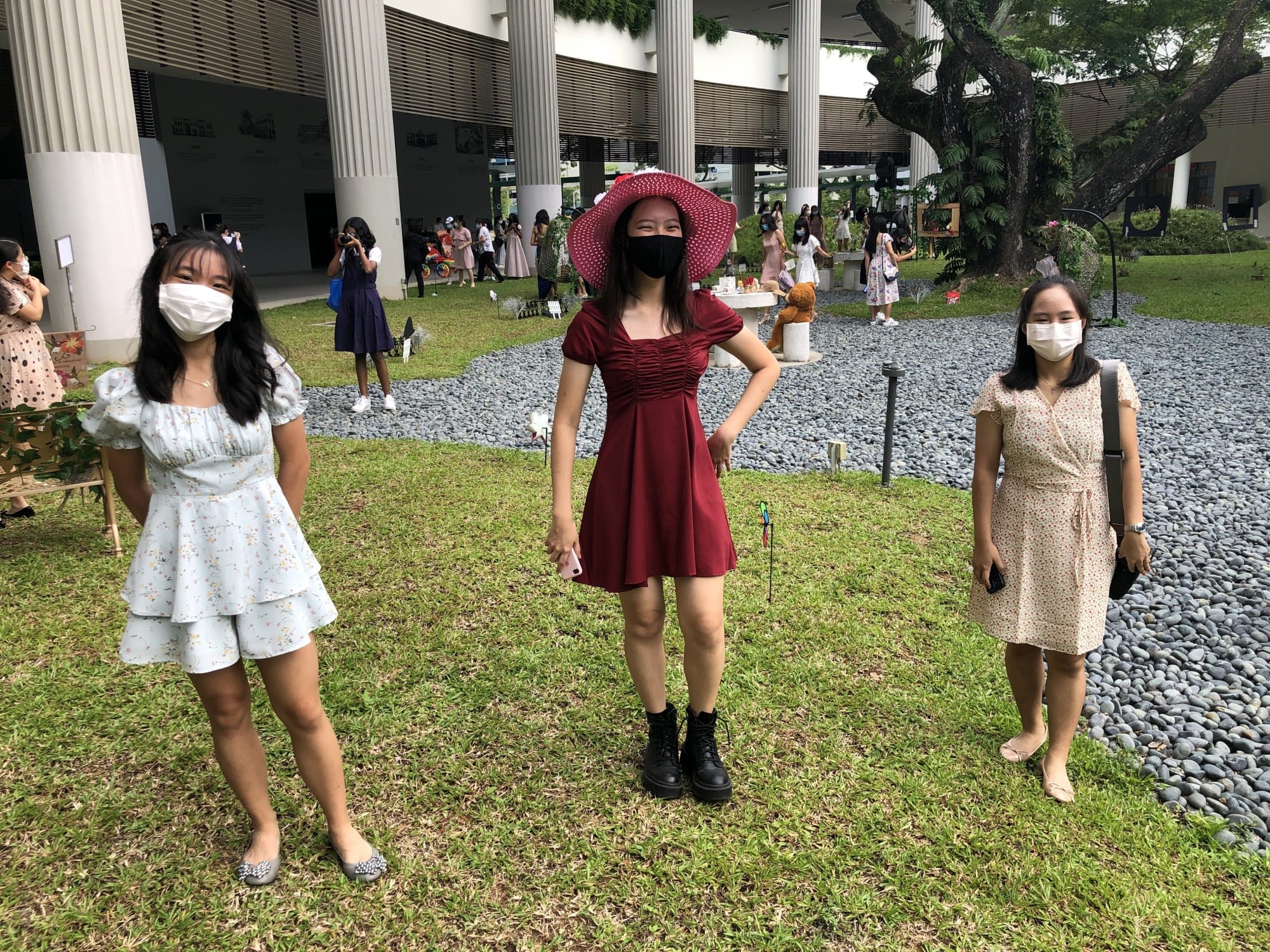 Three young women wearing face masks stand on grass outside a modern building with tall columns.