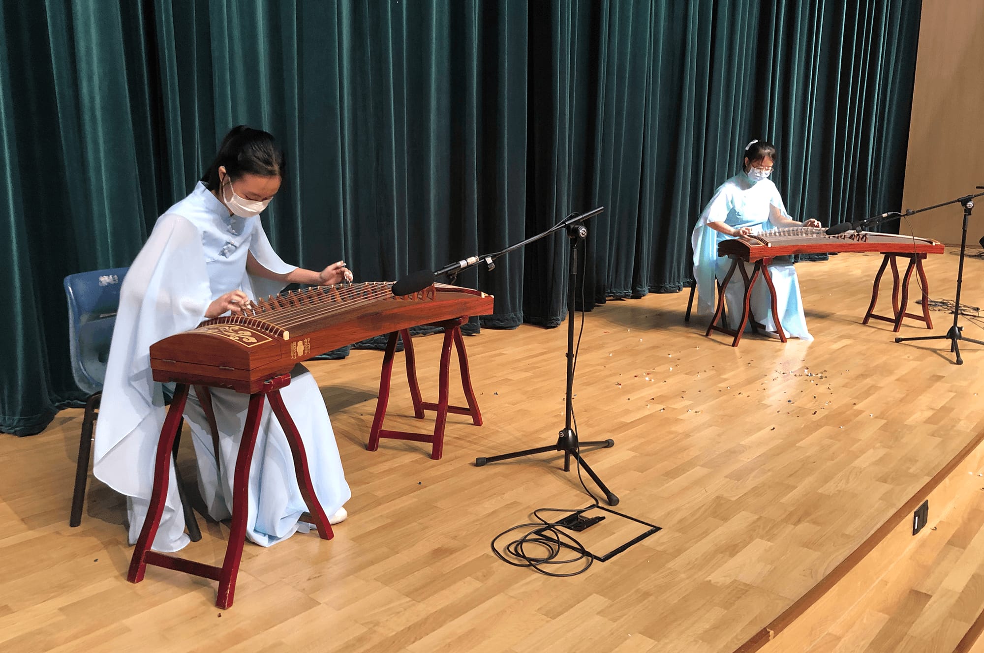 Two masked performers in light blue dresses play guzheng instruments on a wooden stage with a teal curtain backdrop.