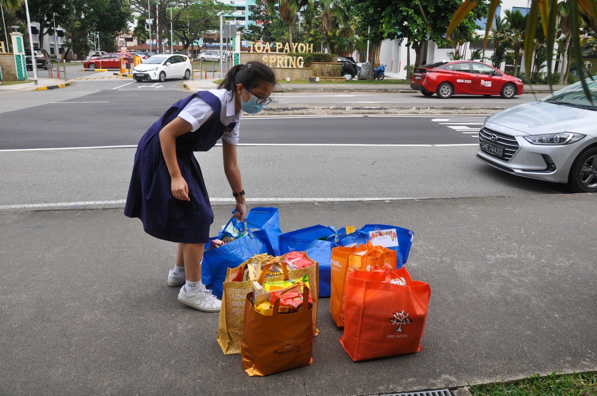 A masked schoolgirl in uniform bends over several colorful reusable bags filled with goods near Toa Payoh Spring.