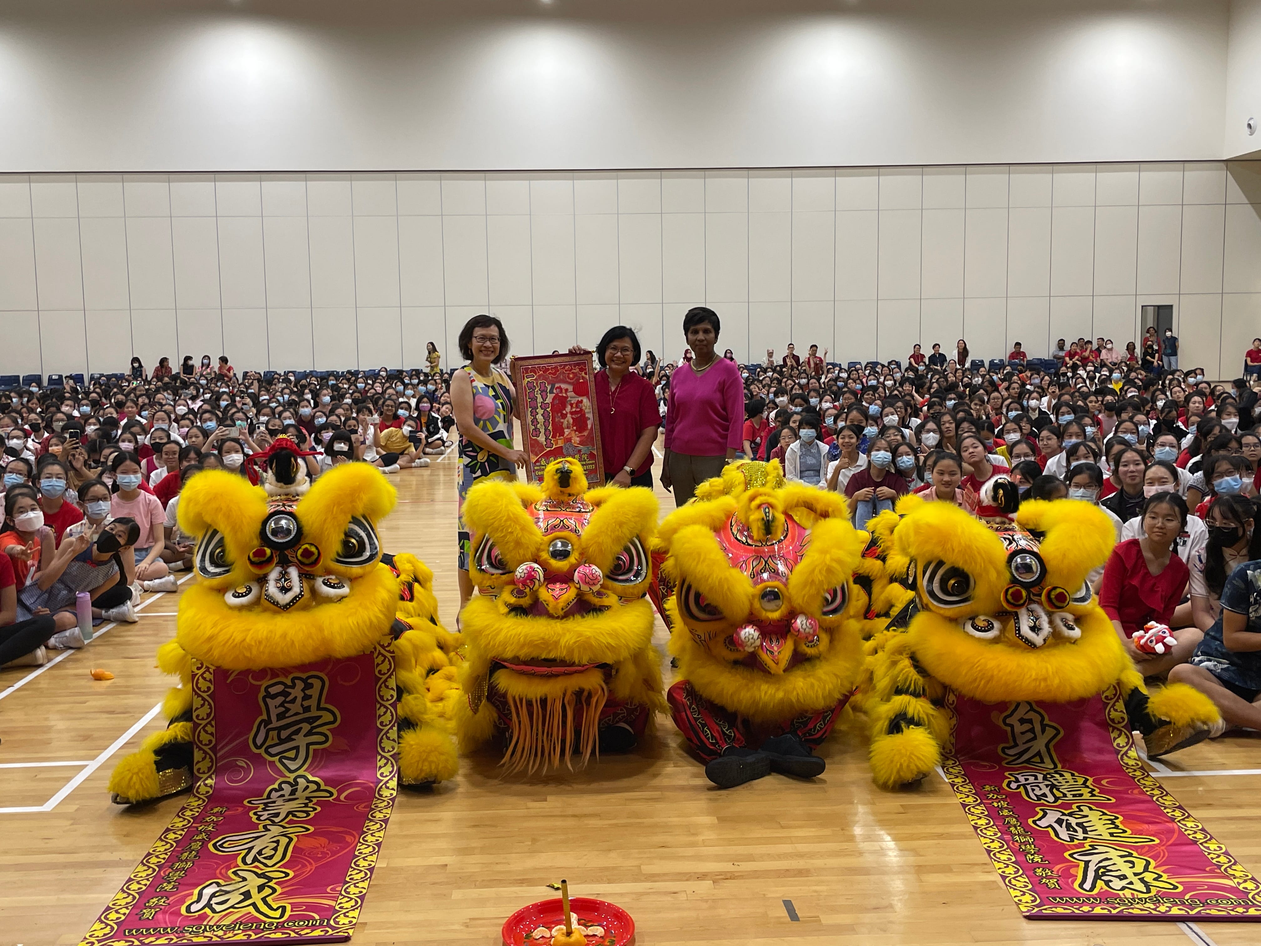 Three adults hold a decorative plaque behind four yellow Chinese lion dance costumes in a packed school gymnasium.