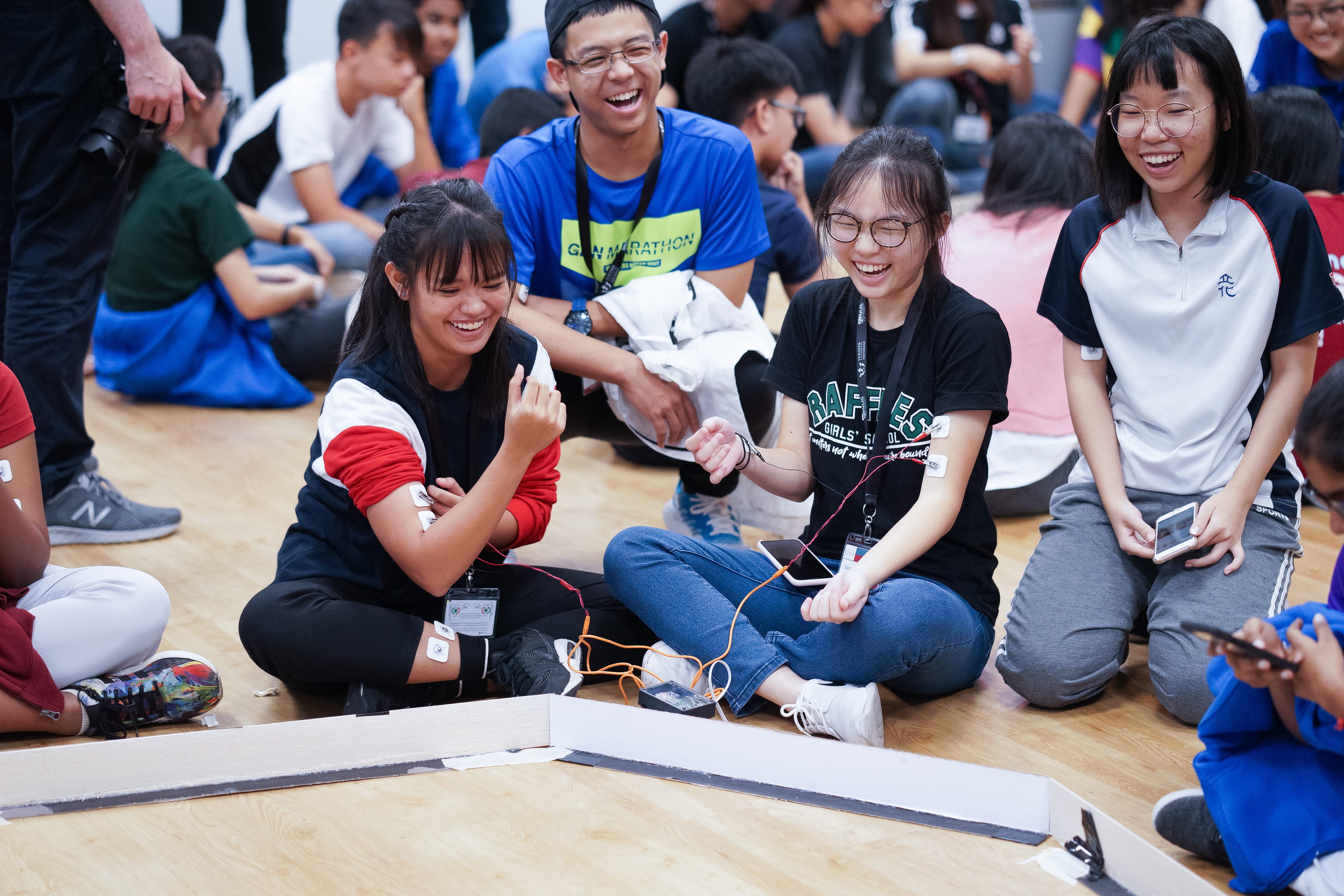 Students sit on a gym floor laughing, with wires and electrodes attached to their arms during an activity.