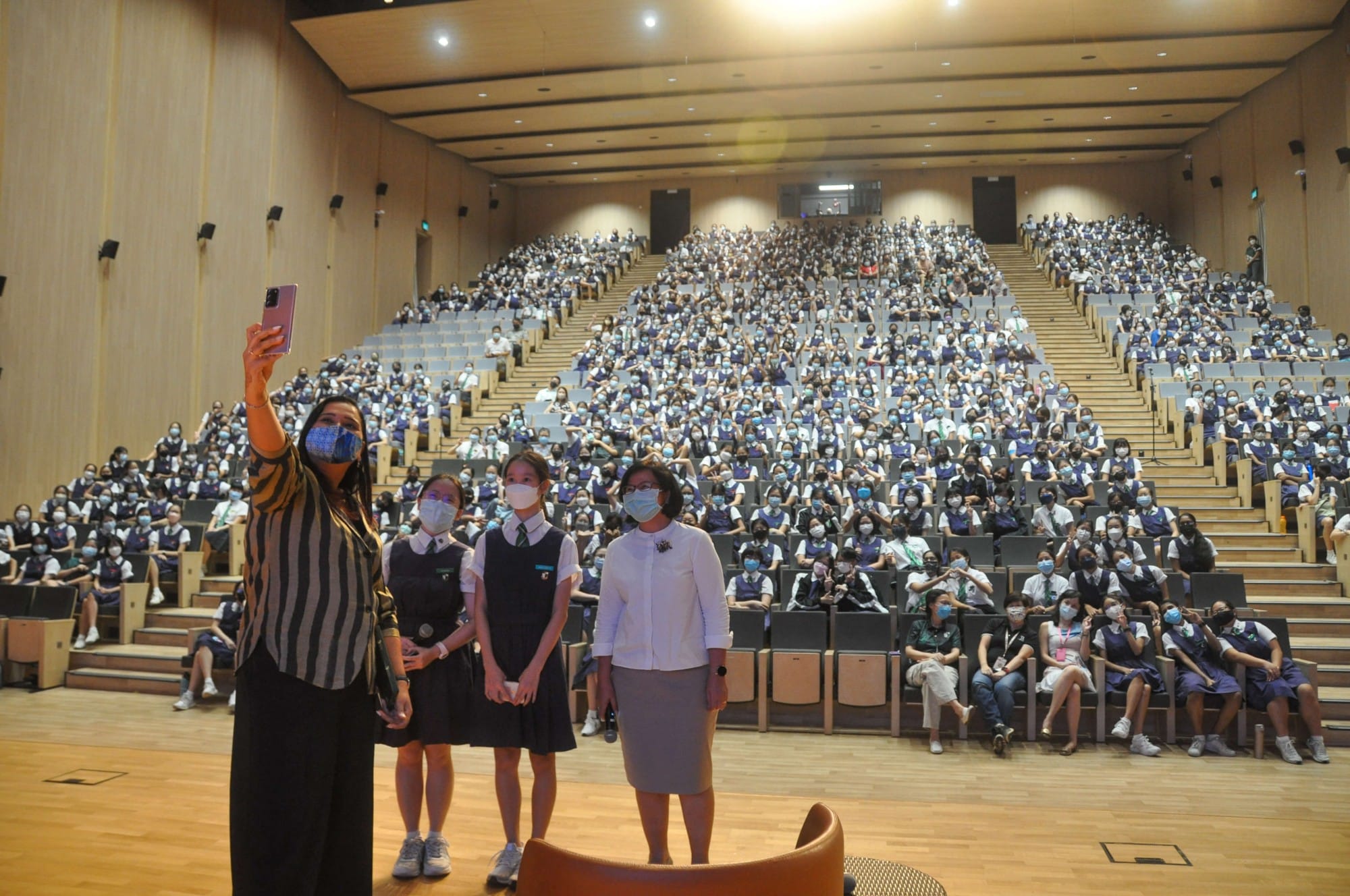 A woman takes a selfie with three others in front of a large auditorium filled with masked students in uniforms.