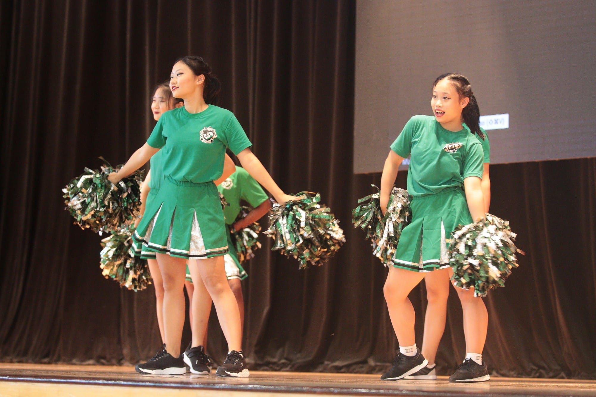 Four cheerleaders in green uniforms perform on stage holding green and silver pom-poms.
