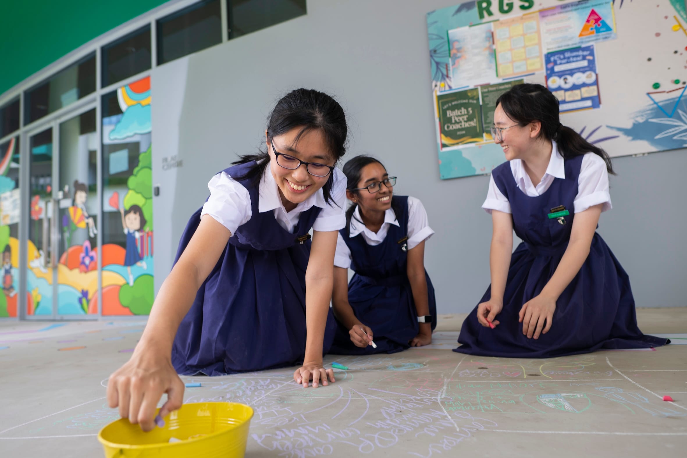 Three schoolgirls in navy uniforms seated on the ground, drawing on the floor with chalks and smiling at each other.