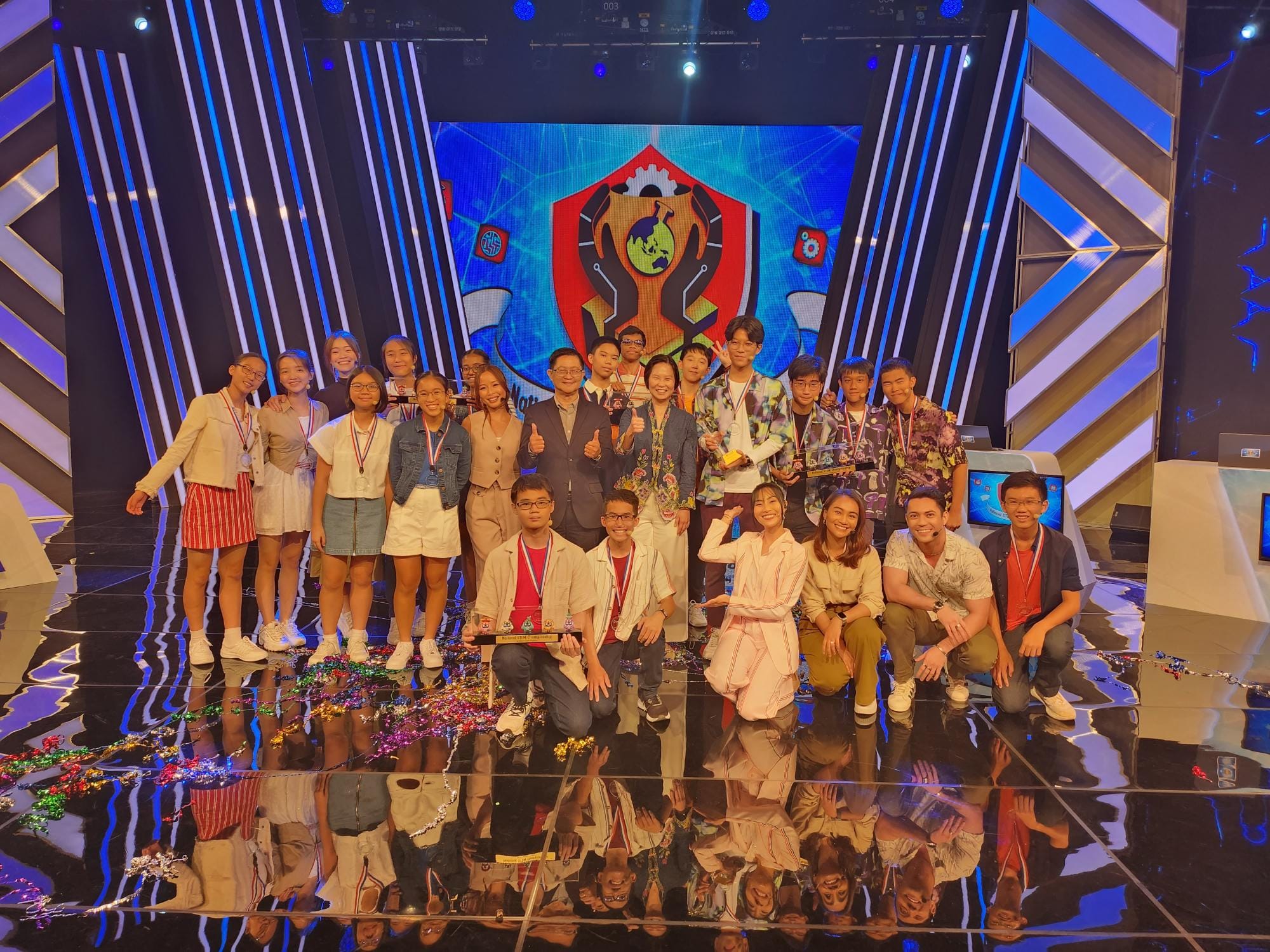 A large group of young people wearing medals pose on a colorful TV competition stage with a shield emblem on screen.