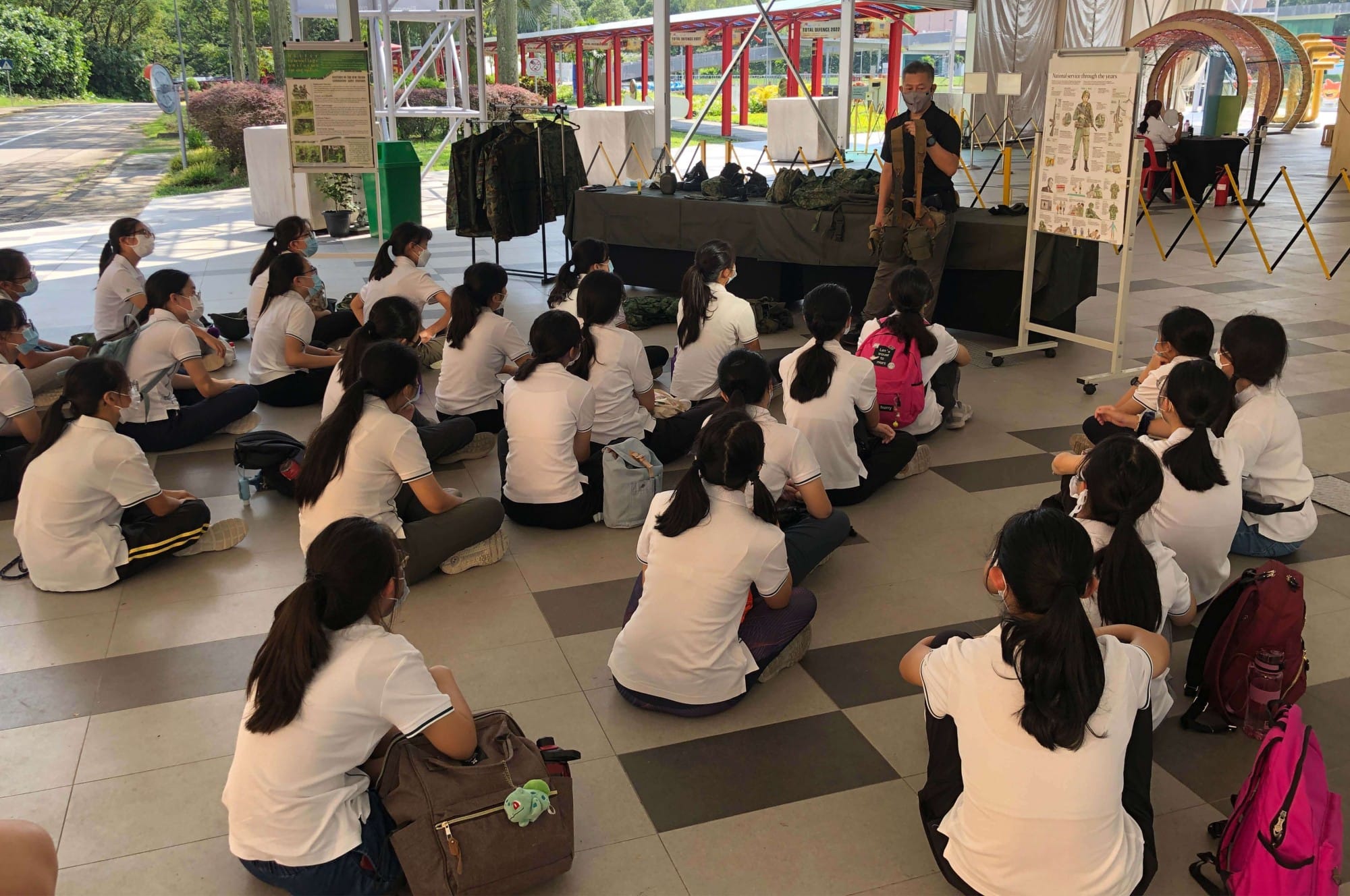 Students in white uniforms sit on the floor listening to a masked instructor presenting near military gear and informational boards.