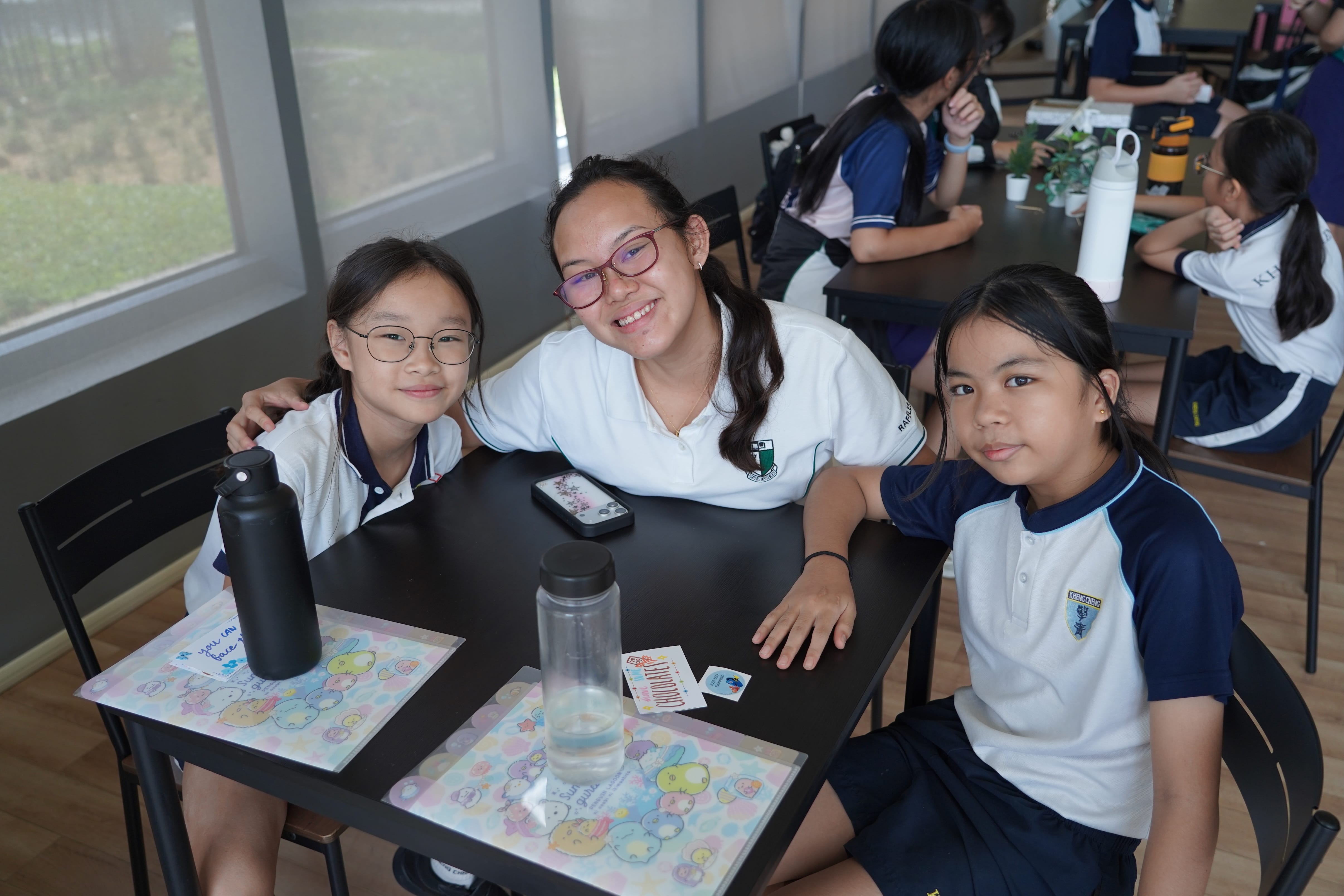 Three smiling students in school uniforms sit together at a desk with water bottles and papers in a classroom.