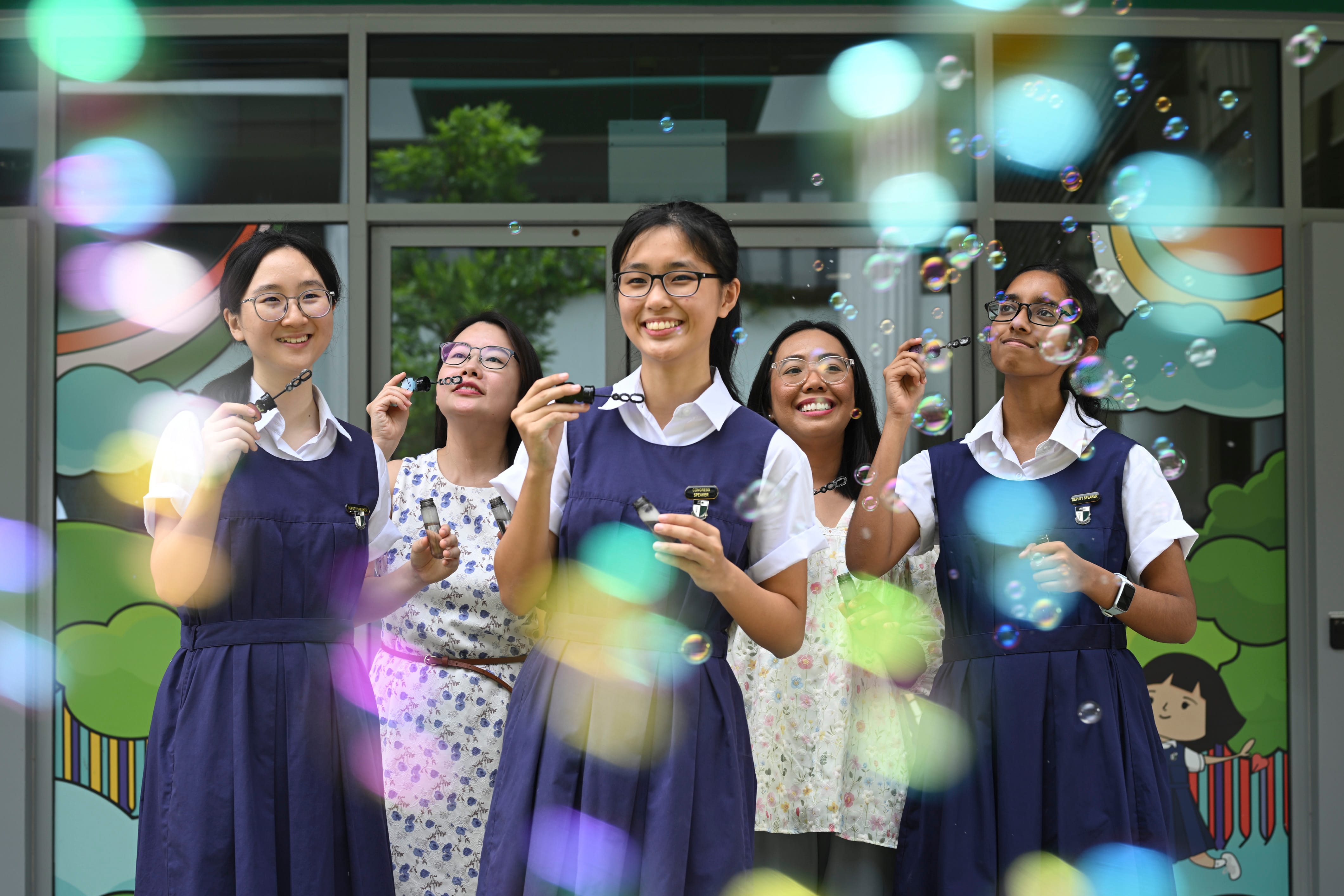 Five smiling young women in navy uniforms blow soap bubbles outdoors near a colorful mural.