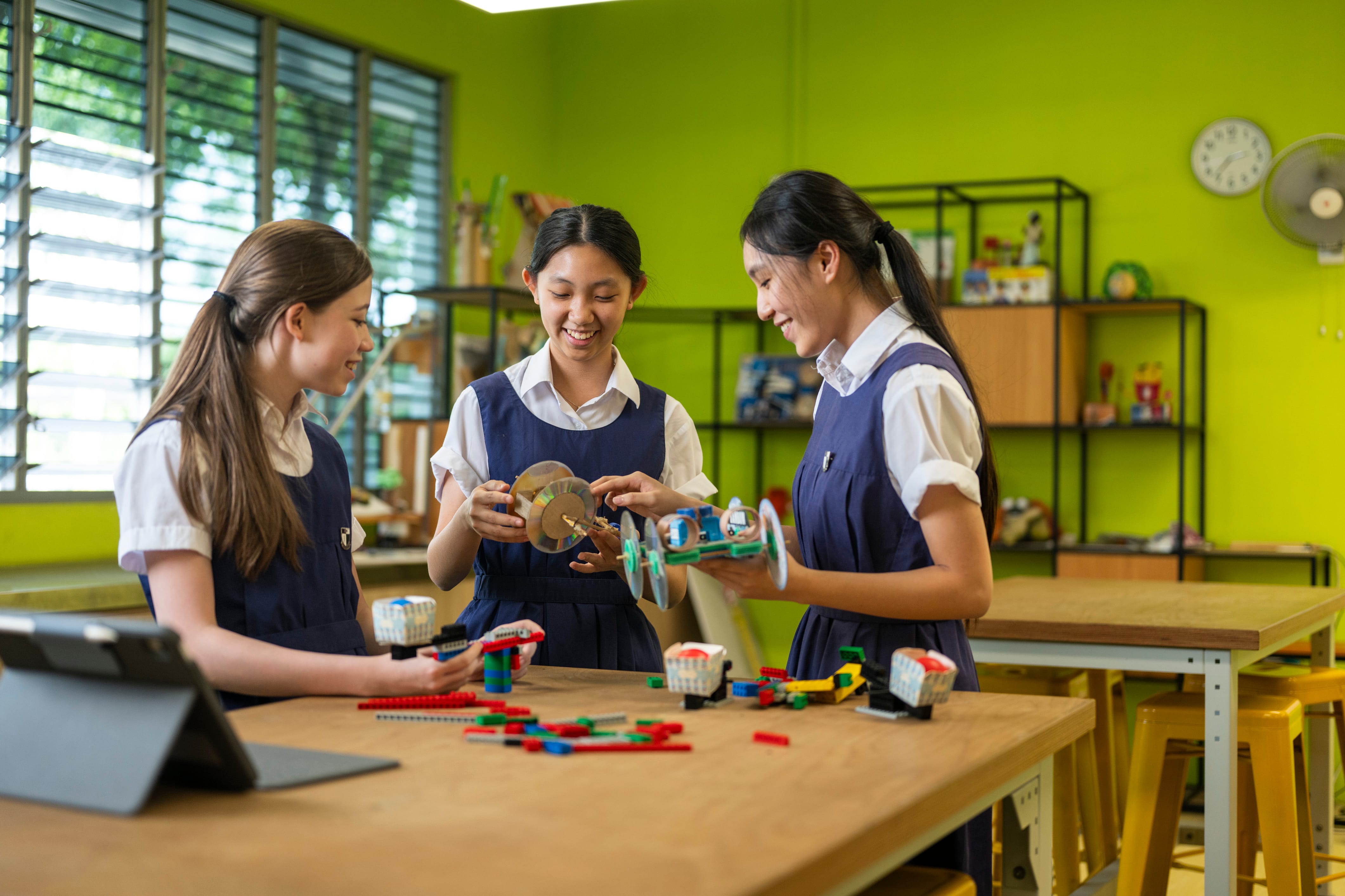 Three smiling schoolgirls in uniforms build wheeled models with colorful bricks on a table in a bright green classroom.