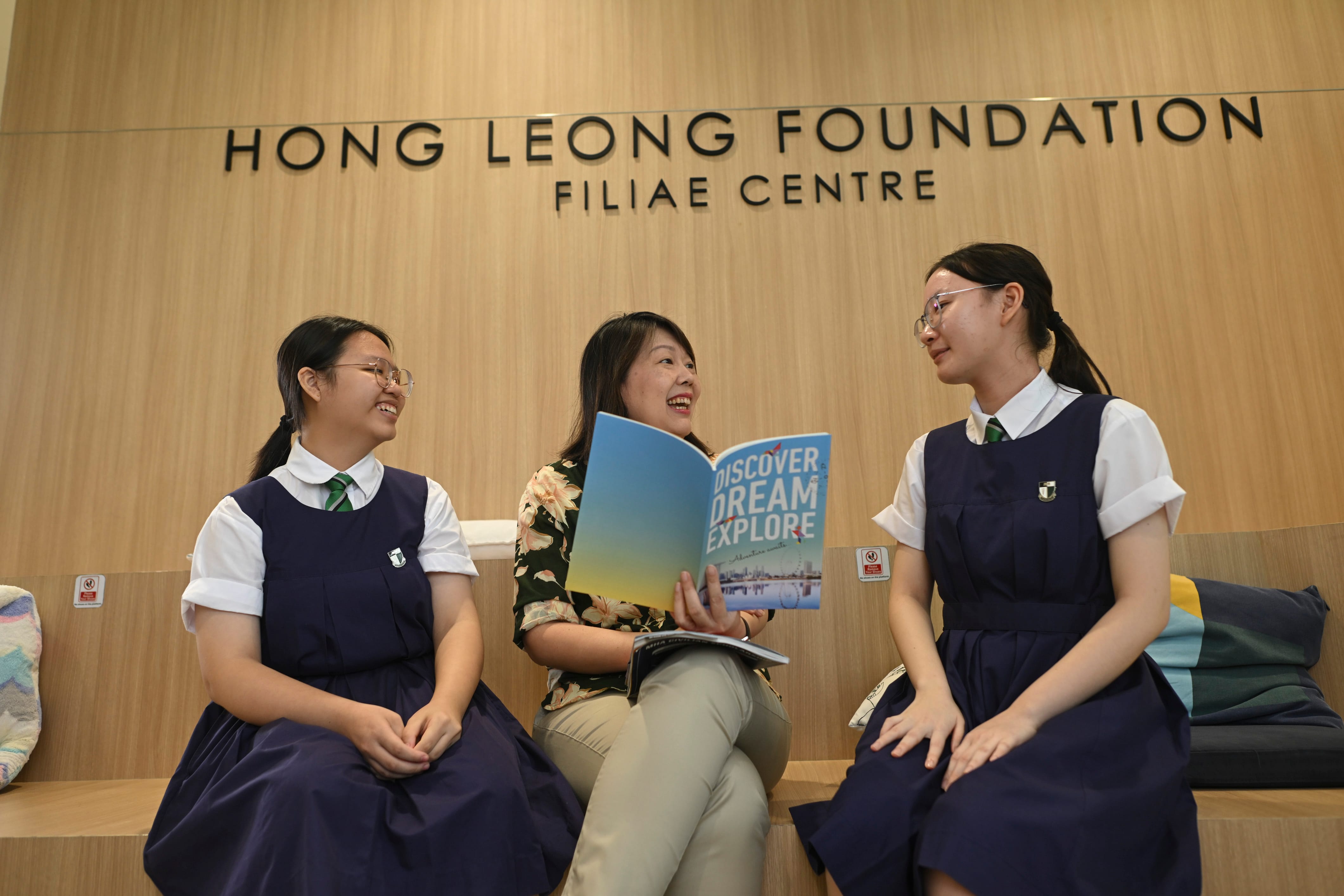Three women sit smiling in the Hong Leong Foundation Filiae Centre, with a teacher holding a Discover Dream Explore booklet.