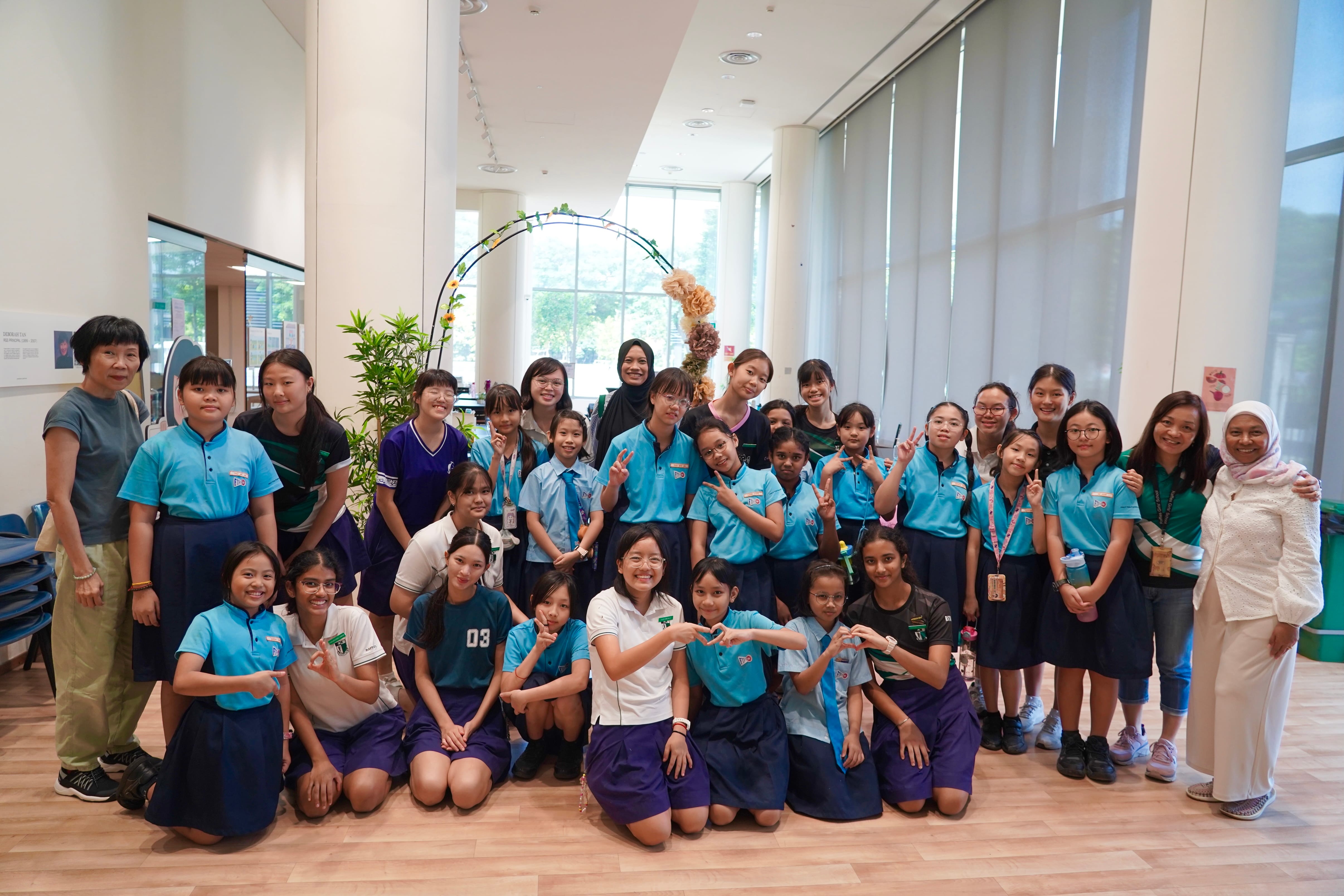 A large group of school girls in blue uniforms pose with adults indoors near a decorative floral arch.