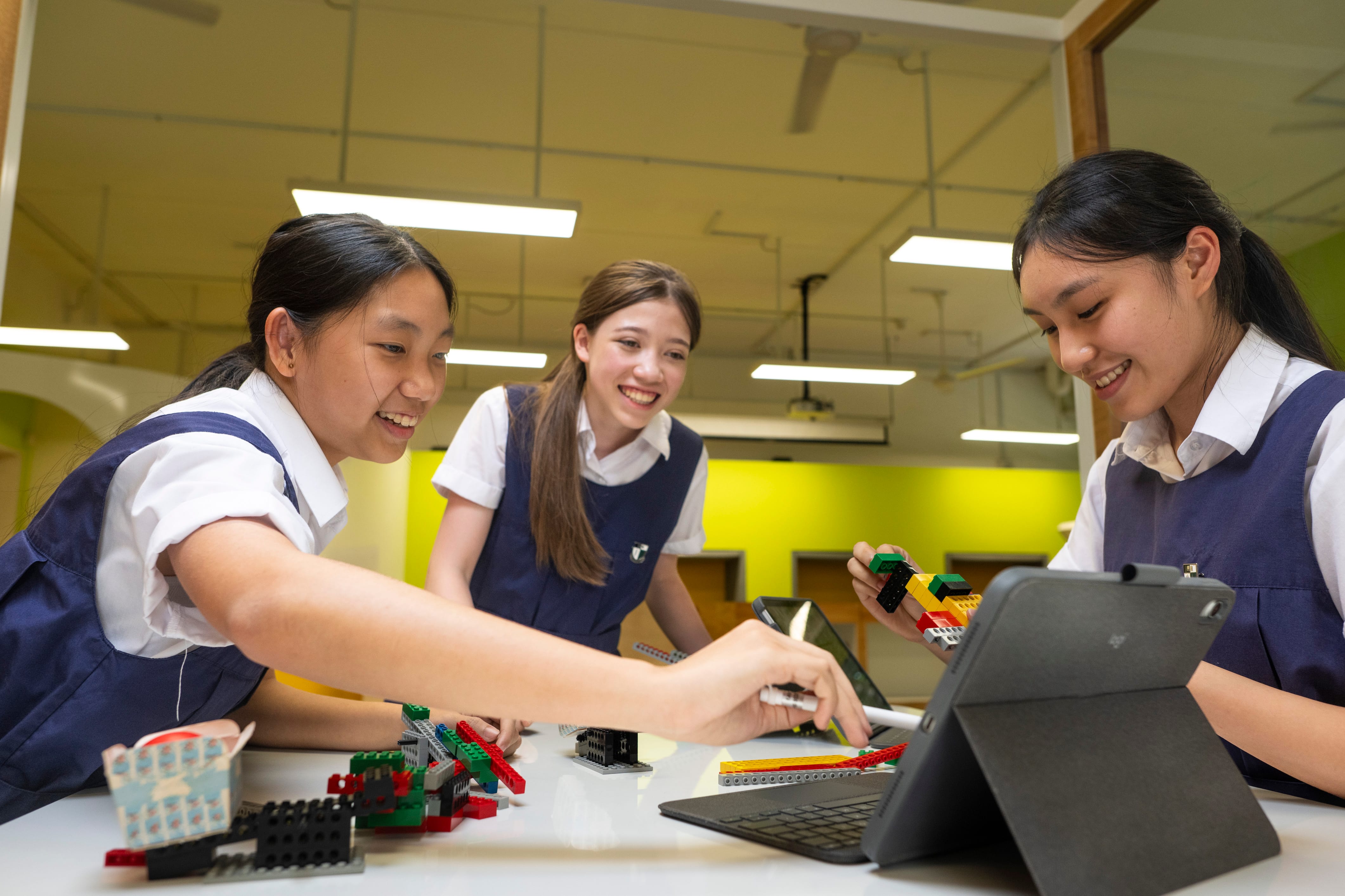 Three smiling schoolgirls in uniforms work together with colorful LEGO bricks and a tablet at a classroom table.
