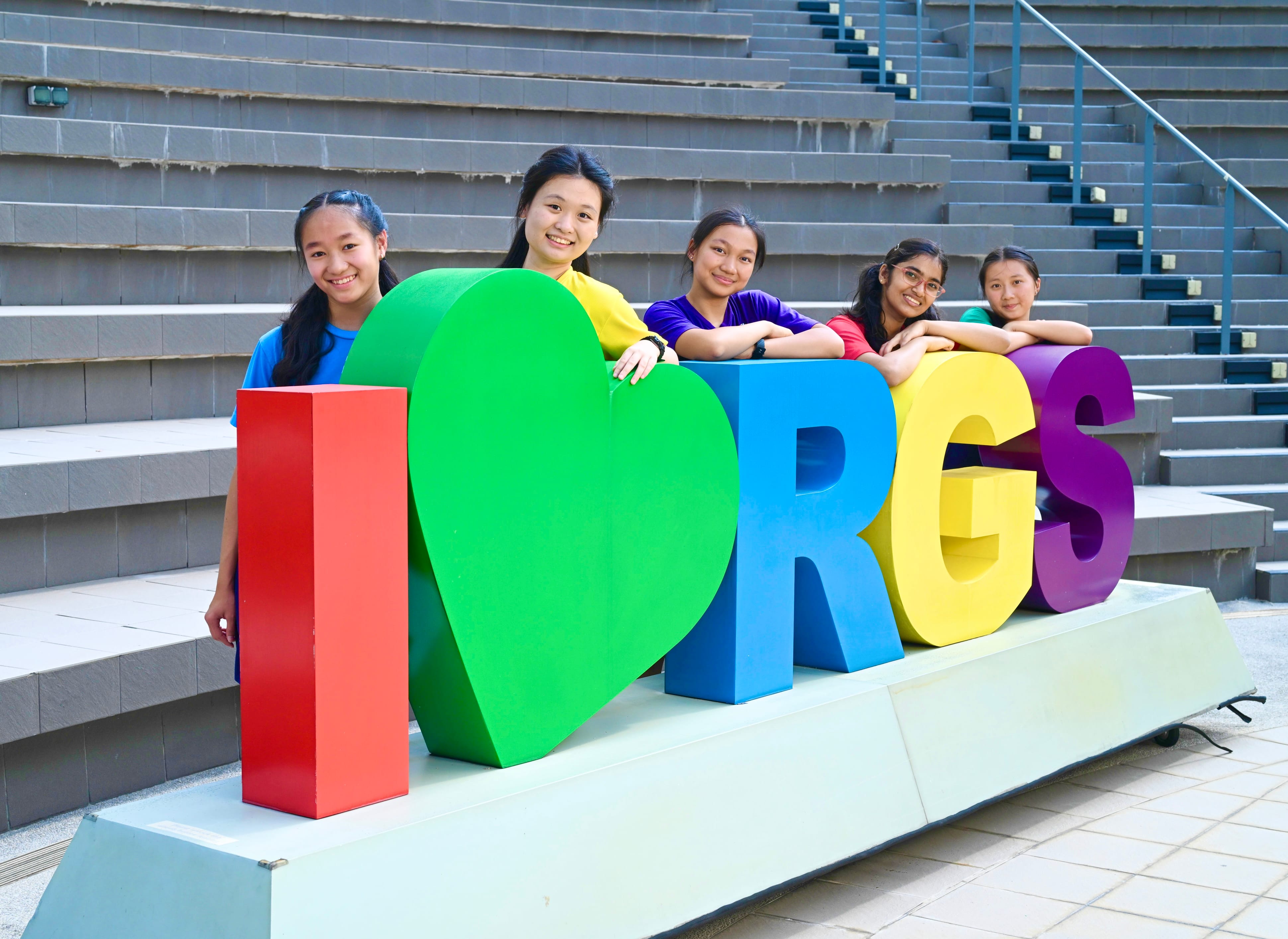 Five smiling girls pose behind large colorful 3D letters spelling "I Love RGS" in front of stone bleachers.