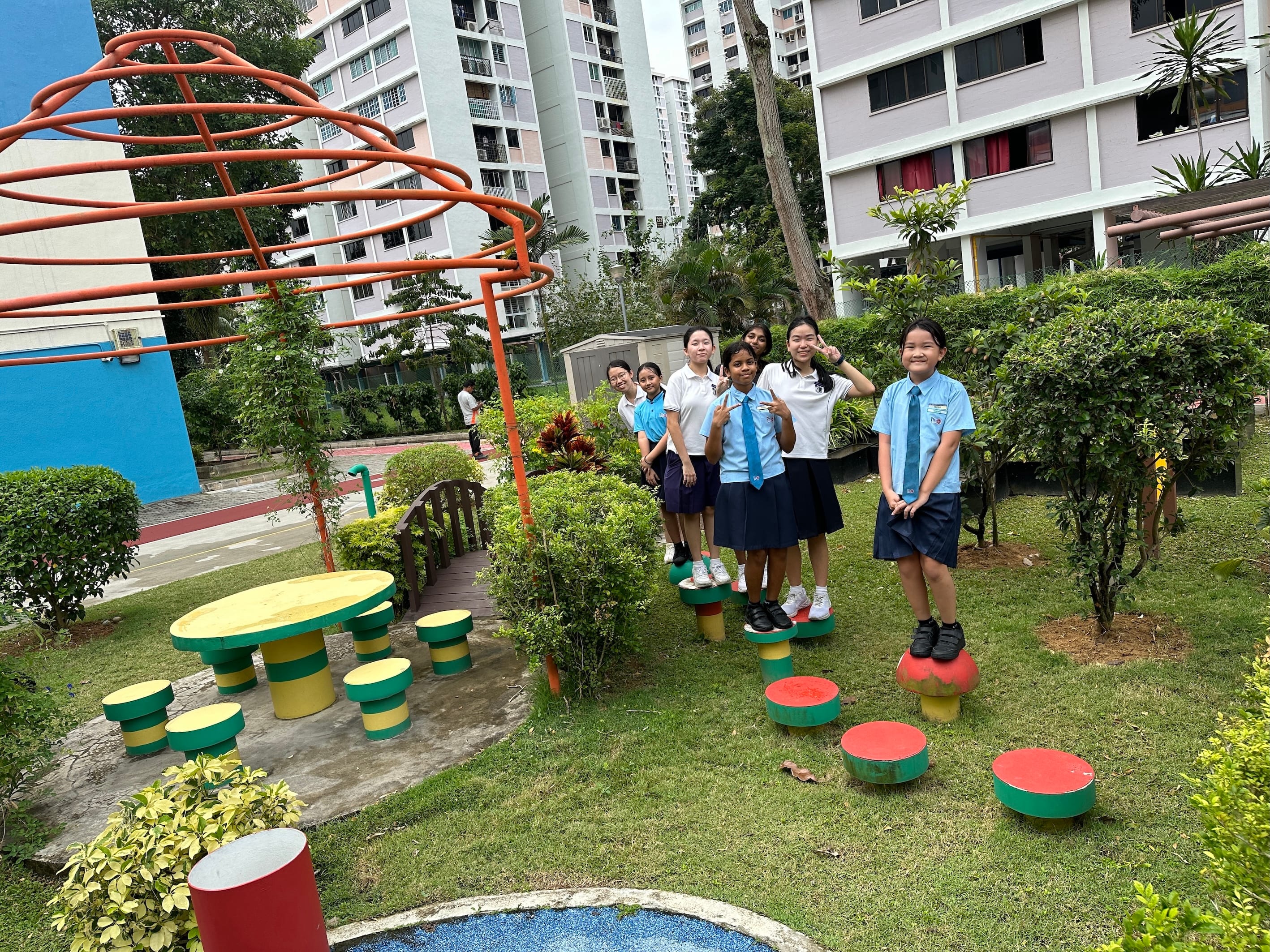 Six schoolchildren in uniforms stand on colorful stepping pods in a garden courtyard with tall apartments behind.