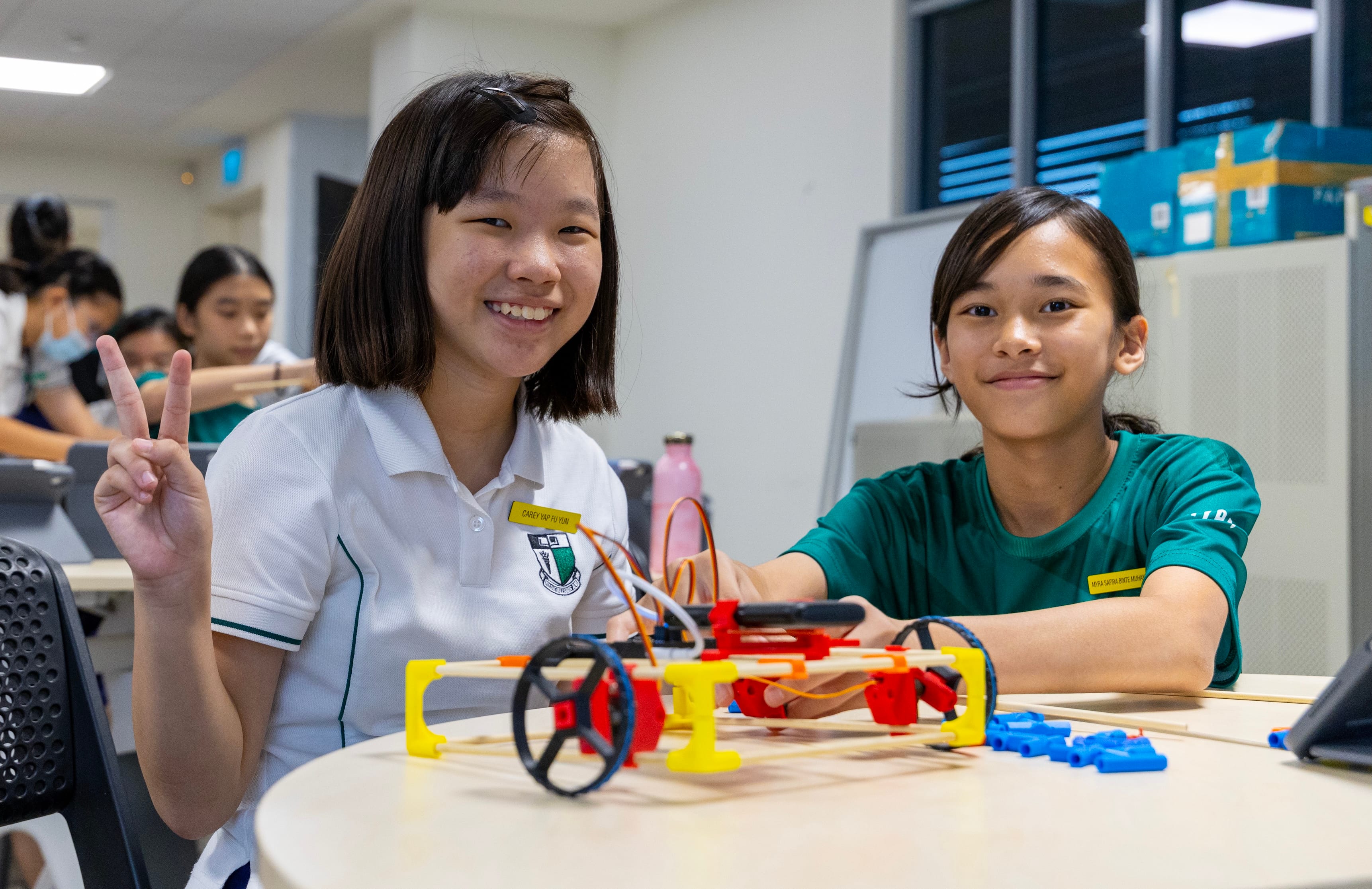 Two smiling schoolgirls sit at a table with a colorful DIY robot vehicle; one flashes a peace sign.