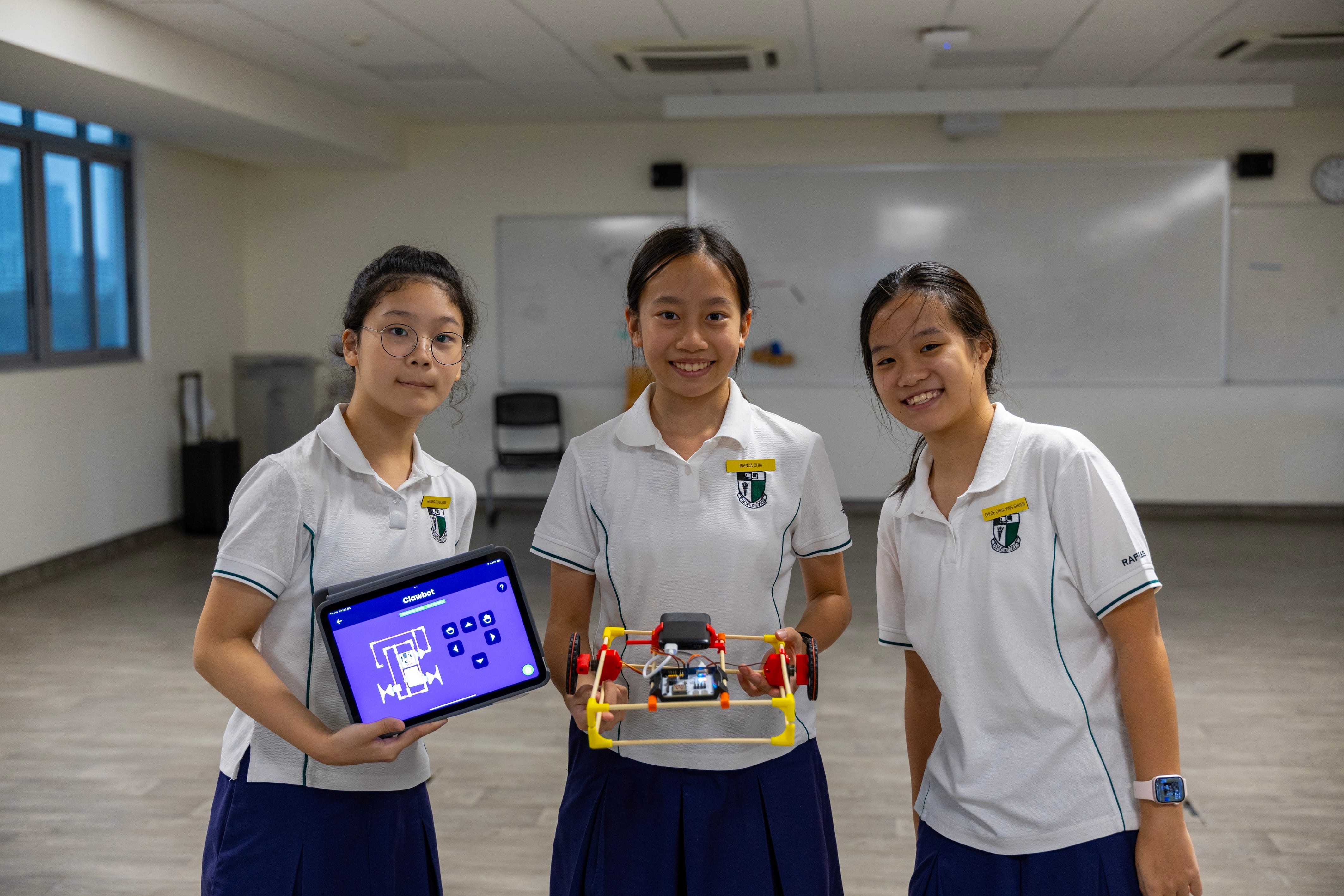 Three schoolgirls in uniforms in a classroom; one holds a tablet, another holds a small robot car.