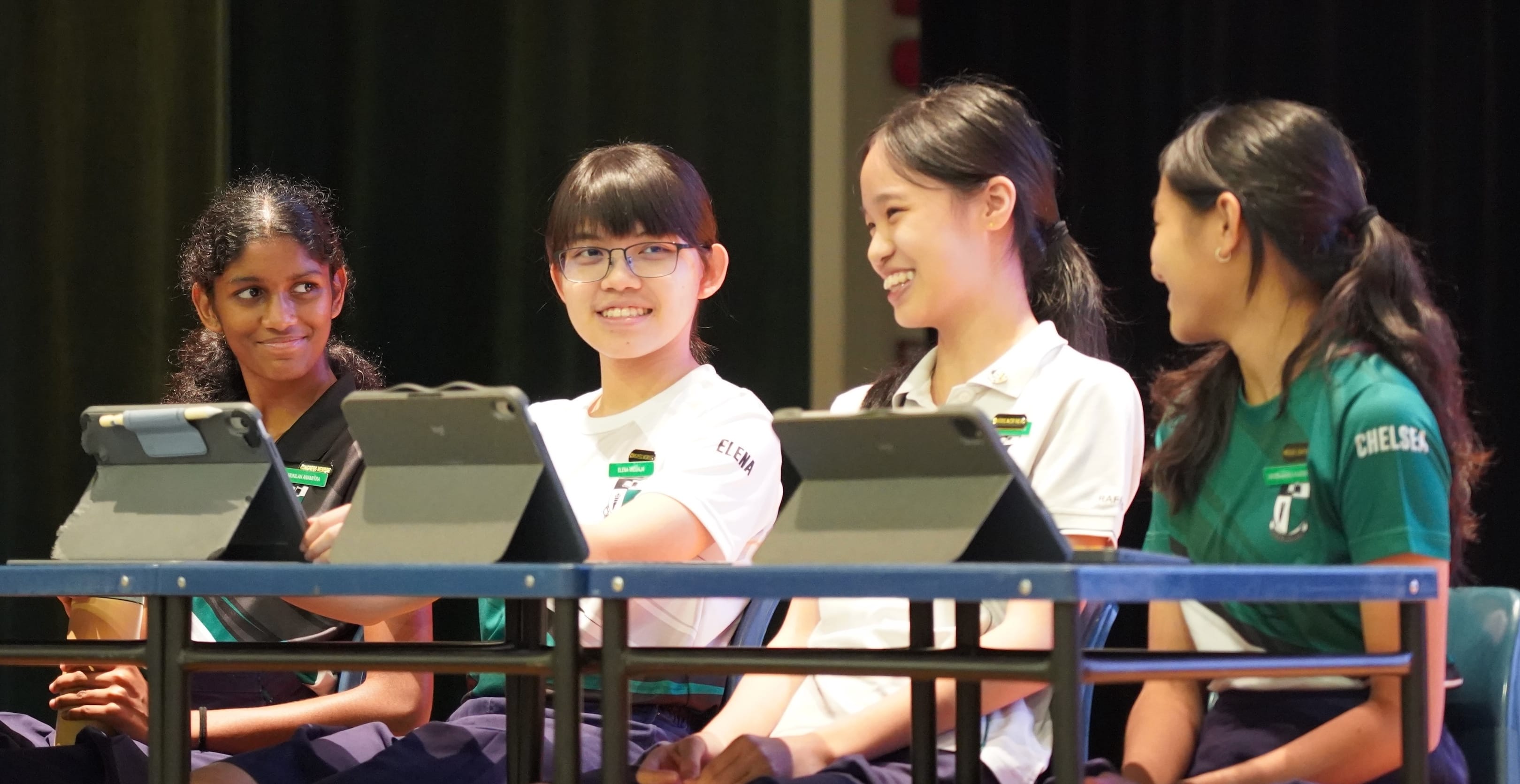 Four smiling schoolgirls in uniforms sit at desks with tablets, wearing name tags, in a darkened hall.