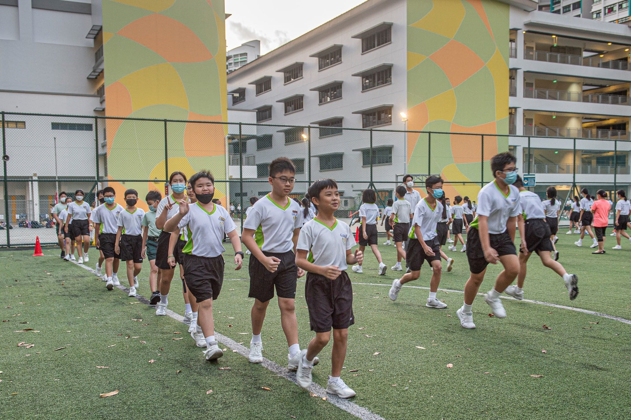 Students in white and black uniforms wearing face masks walk and jog on a green sports field near school buildings.