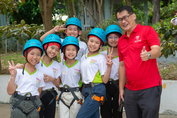 Six children in blue helmets and harnesses pose with a smiling adult man in a red shirt giving a thumbs up.
