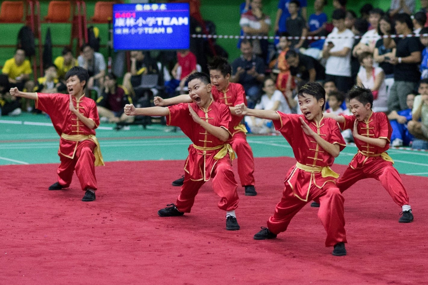 Five children in red and gold uniforms perform a synchronized wushu routine on a red mat before a crowd.