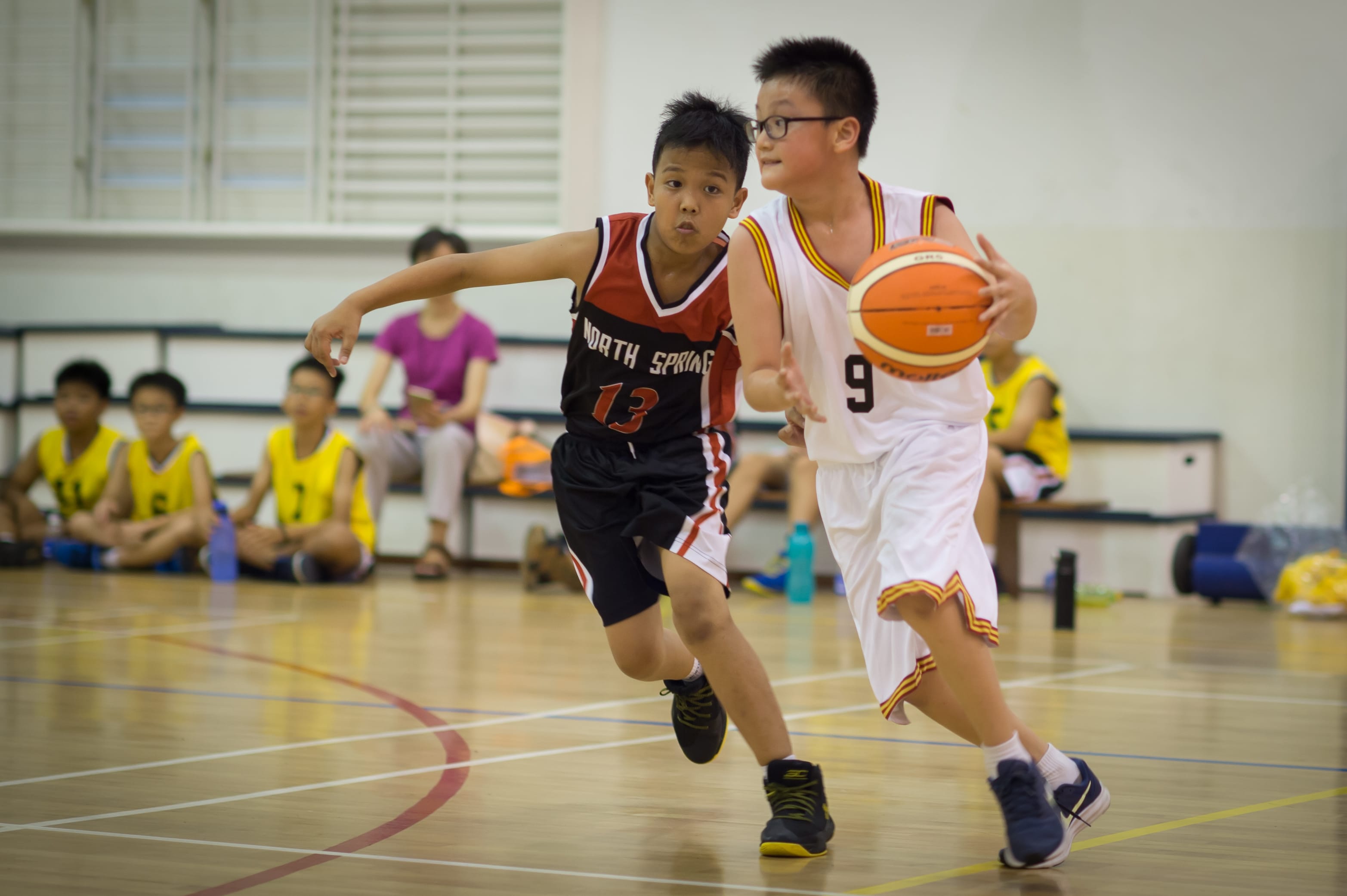 Two boys play basketball indoors, one in white jersey number 9 dribbling while number 13 in black defends.