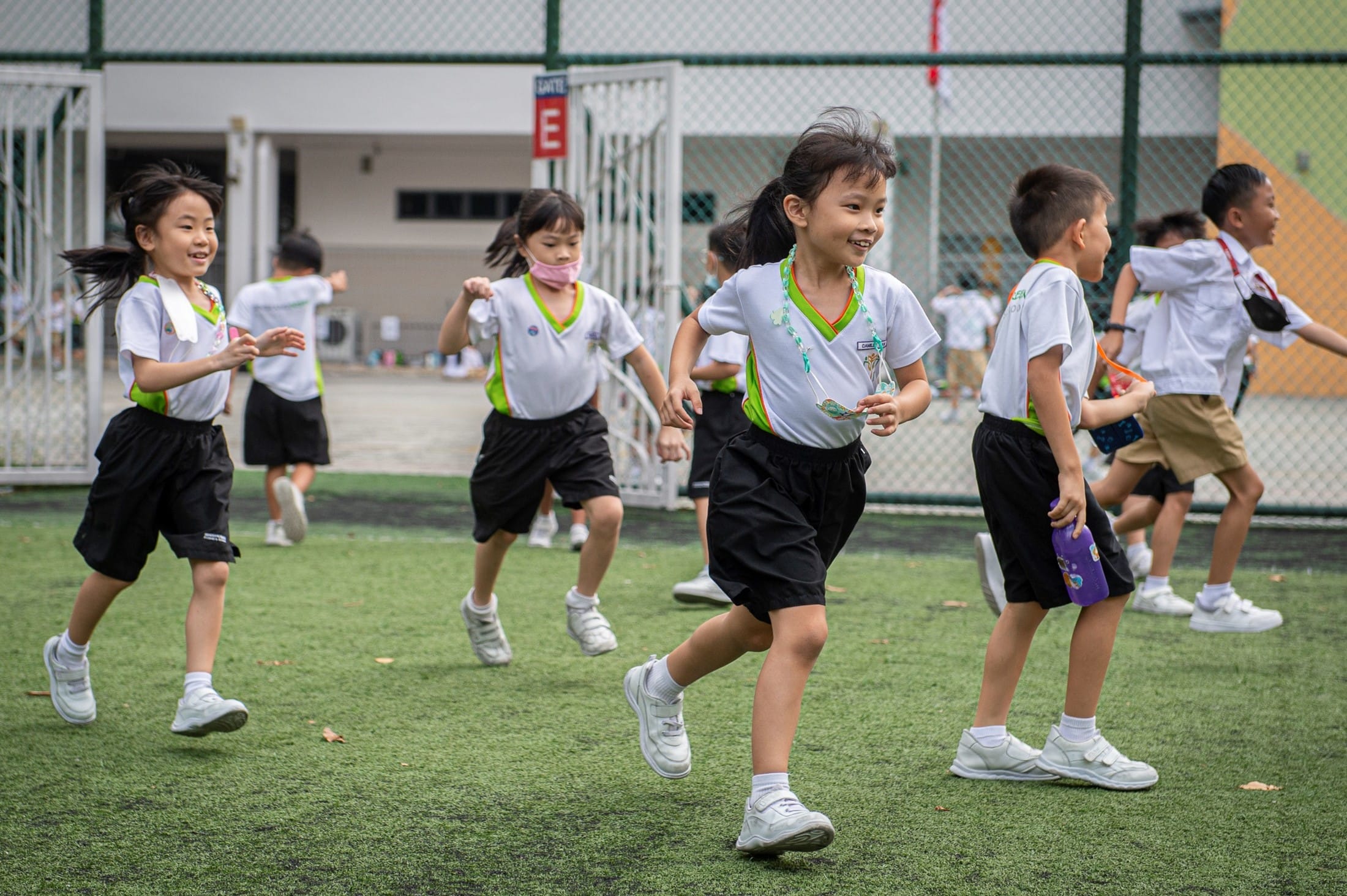Young children in white and green school uniforms run and play on a green turf field outdoors.