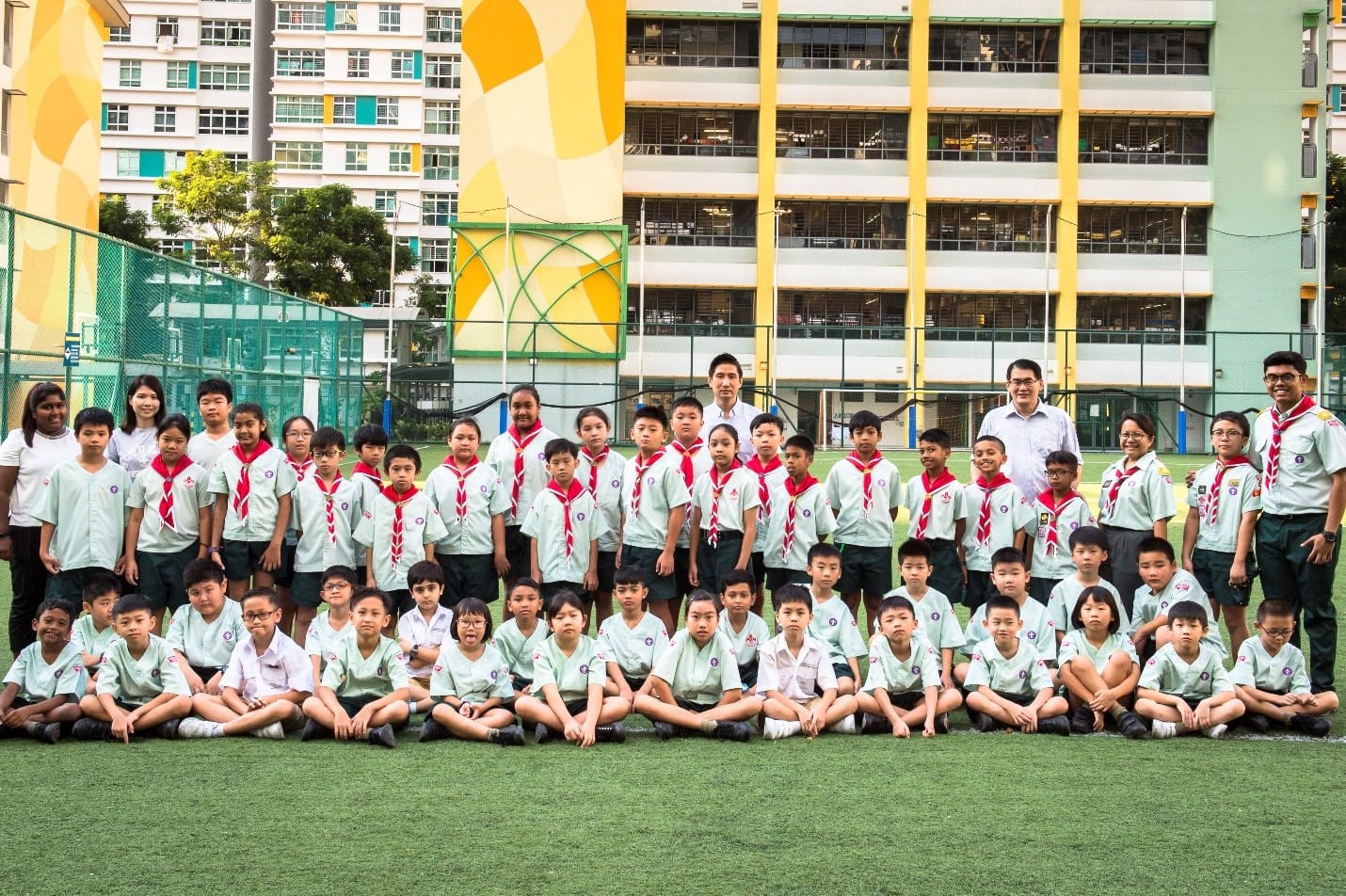 A large group of children in light green scout uniforms with red scarves pose on a field with adults behind them.