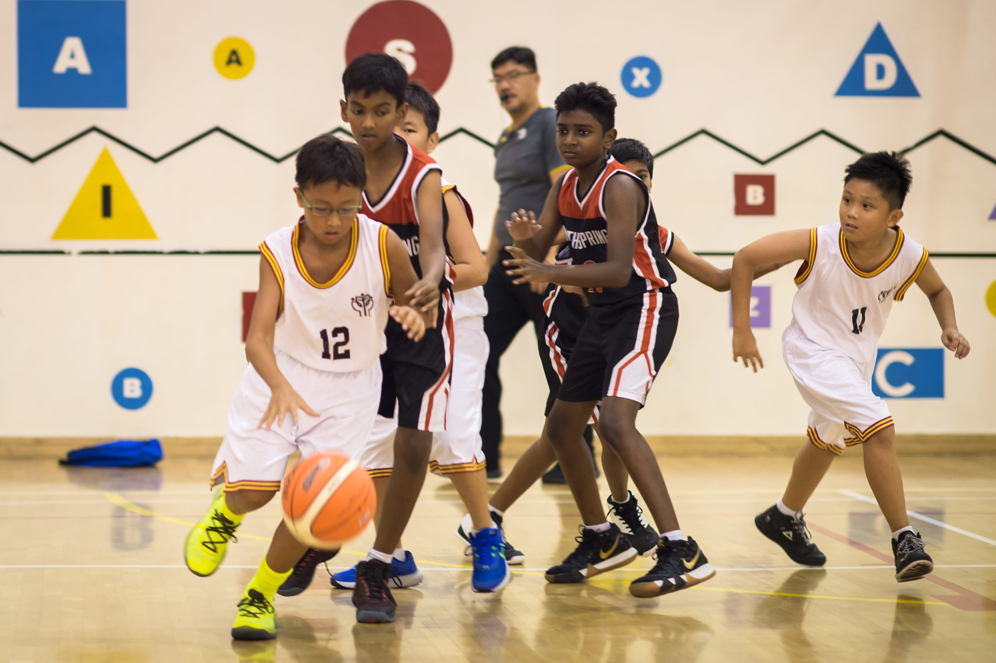 Young boys in white and black jerseys play basketball on an indoor court decorated with colorful letters and shapes.