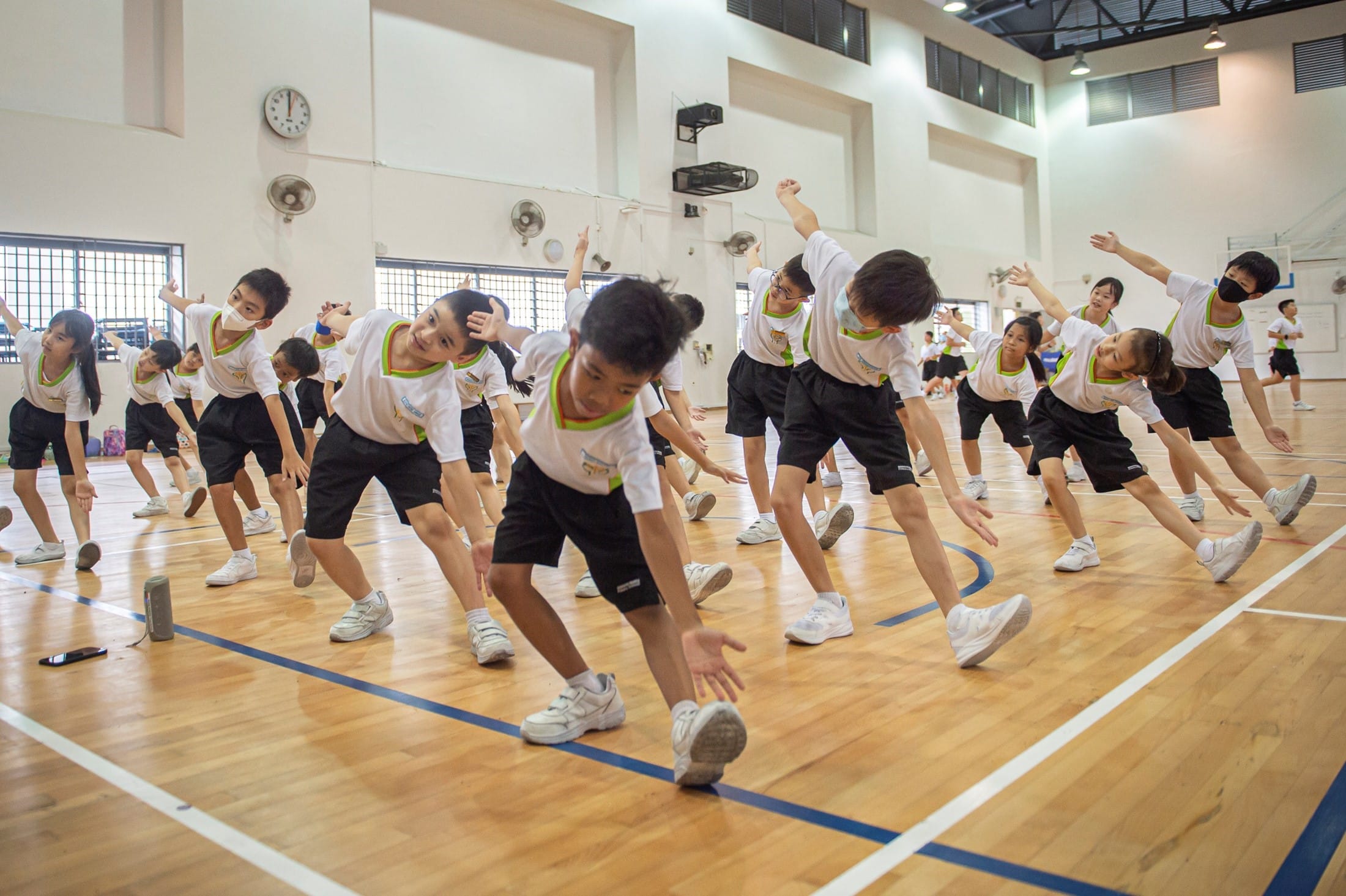 Children in white shirts and black shorts do stretching exercises together on a wooden gymnasium floor.