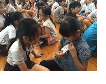 Two children sit on a wooden floor looking at a small card, surrounded by many other children seated nearby.